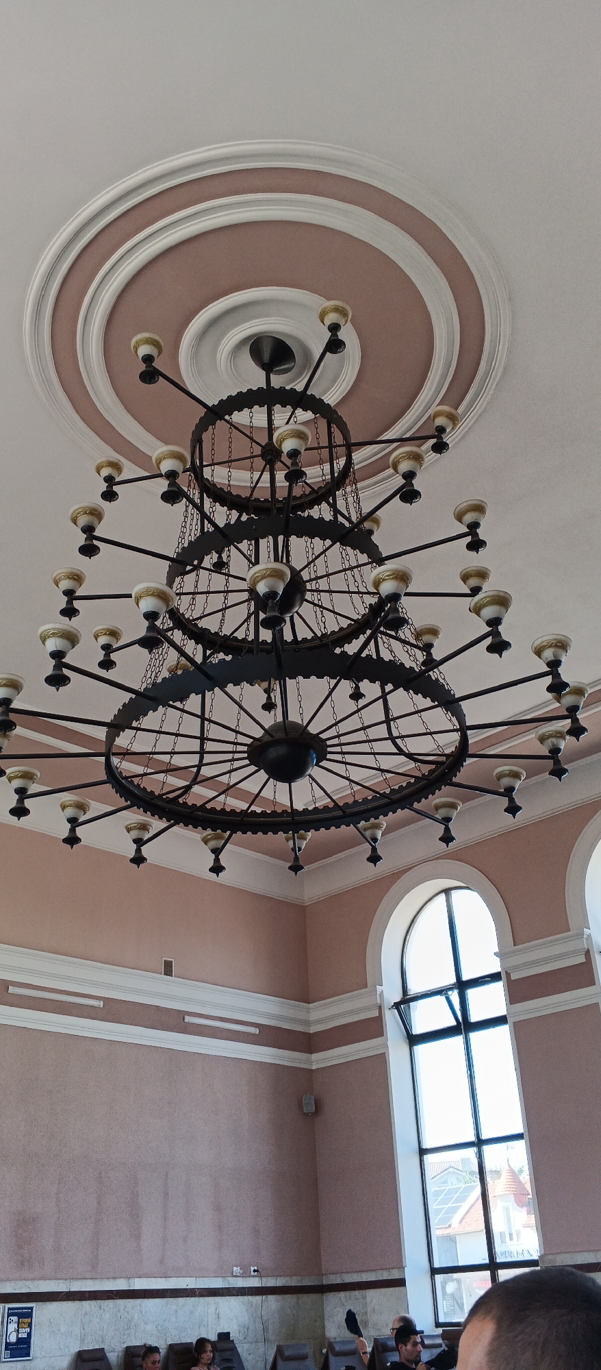 The image shows a large, multi-tiered chandelier hanging from a decorated ceiling in what appears to be a public building. The chandelier is made of dark metal with multiple arms and hanging chains, supporting many light fixtures. The ceiling has concentric circles of molding, with the central area painted a dusty pink.
The room has pale pink walls with white trim. In the background, a large arched window with multiple panes looks out onto buildings and rooftops, suggesting an urban setting. Below the window, the lower part of the wall has a different texture and color.
In the foreground, several people are visible, seated or standing. Some appear to be in a waiting area or a lobby. One person in the bottom right is mostly obscured, with their head in the frame. Another person is holding up a dark object, possibly a hat or a bag.
This image was taken in the Mukachevo railway station in Mukachevo, Ukraine.