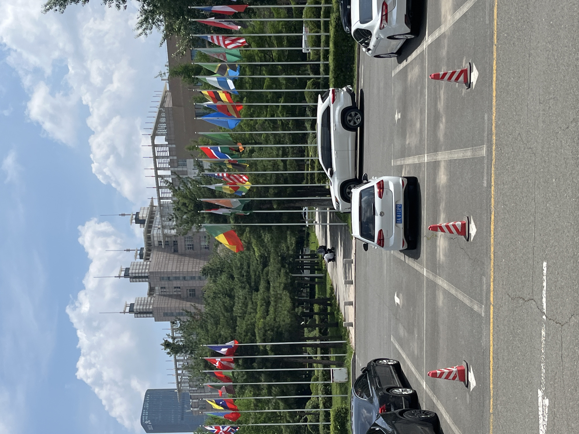 The image captures a bright, sunny day in Changchun City, China, likely at an international or governmental complex, given the prominent display of numerous national flags. In the foreground, a paved parking lot is visible with several cars parked and in motion. Two white cars and one black car are clearly identifiable. Orange and white striped traffic cones are placed on the ground, indicating parking spaces or traffic direction.
Beyond the parking lot, a row of white flagpoles stands tall, each adorned with a different national flag, fluttering in the breeze. These flags create a festive and international atmosphere. Behind the flags, lush green trees form a natural backdrop.
In the middle ground, a paved pathway leads towards a large, multi-story building complex. The architecture of the buildings appears modern and somewhat imposing, with a blend of concrete and glass structures. One particularly tall, dark skyscraper is visible in the distance on the left. A person is seen pushing a stroller on the pathway, adding a human element to the scene.
The sky is a clear blue, dotted with fluffy white clouds, typical of a pleasant summer day. The overall impression is one of a well-maintained, official-looking area, possibly a venue for diplomatic events or international organizations. The presence of cars, flags, and modern architecture suggests an urban setting within Changchun City, China.