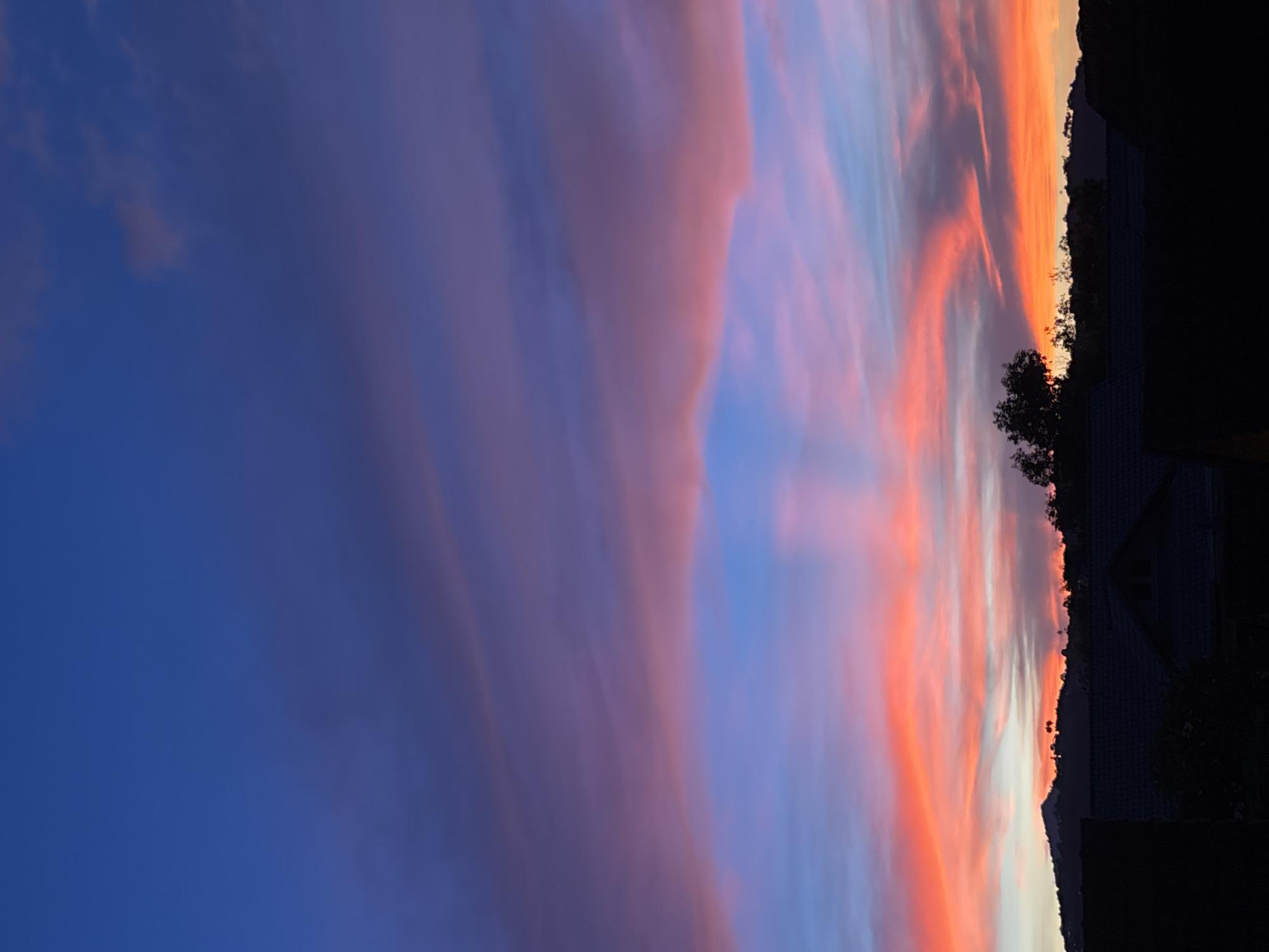 The image shows a sunset sky over บ้านบางรักษ์, Thailand. The sky transitions from deep blue on the left to hues of pink, purple, orange, and light blue towards the right. The silhouettes of trees and rooftops are visible against the bright horizon, indicating the presence of residential buildings. The photograph is taken from a low angle, emphasizing the expansive sky.