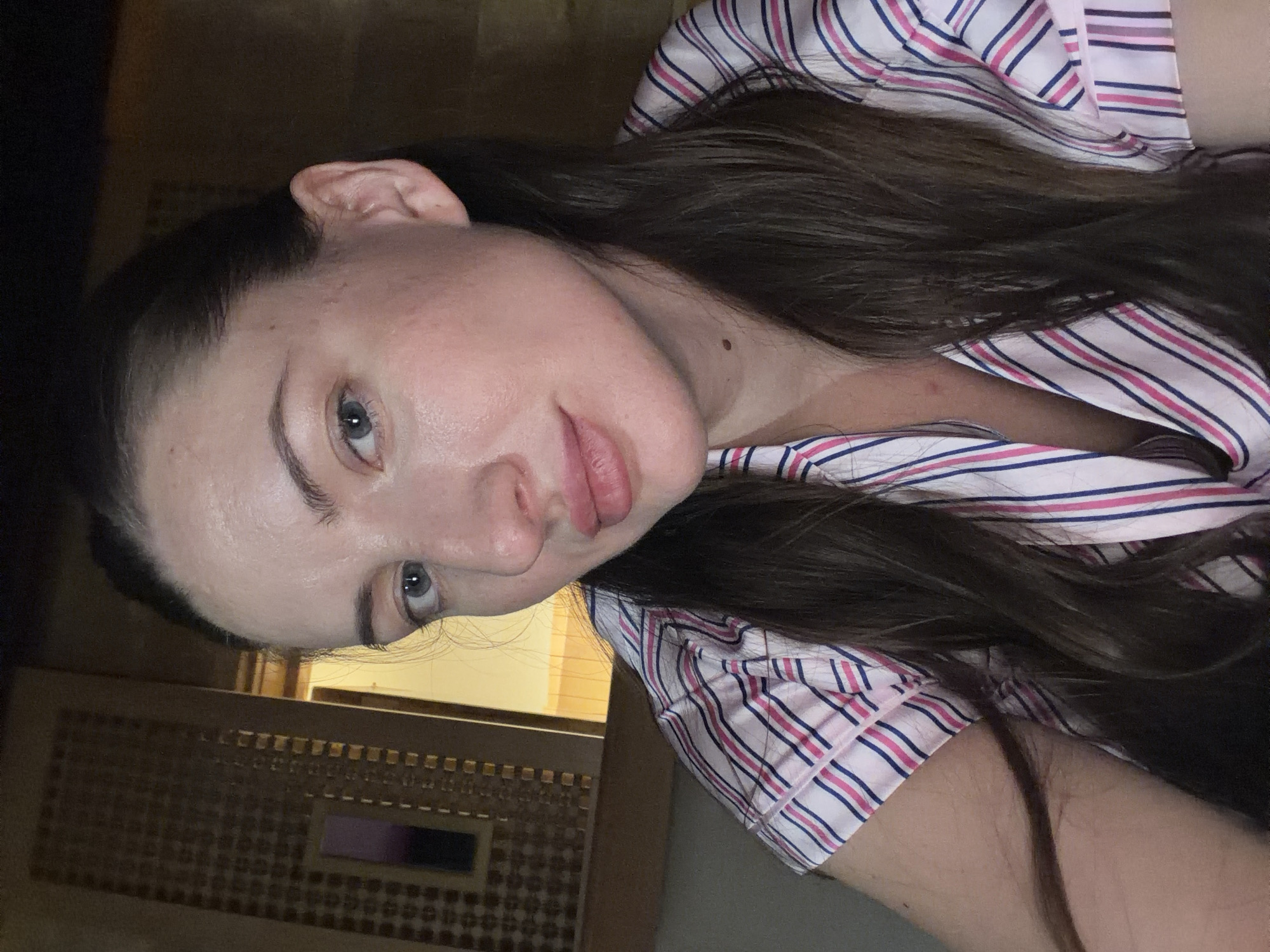 A medium close-up image shows a woman's face against a blurred background that suggests an indoor setting. The woman has fair skin with some redness and appears to be looking directly at the camera. She has light blue eyes, a prominent nose, and full lips. Her dark brown hair is pulled back, with some strands falling around her face and shoulders. She is wearing a pink, white, and blue striped pajama top. The background features a wall with a patterned screen and a hint of light coming from a doorway or window. The image's lighting is somewhat dim, creating a soft glow on the woman's face. There is no indication of the city of Dahab, Egypt, within the image itself.