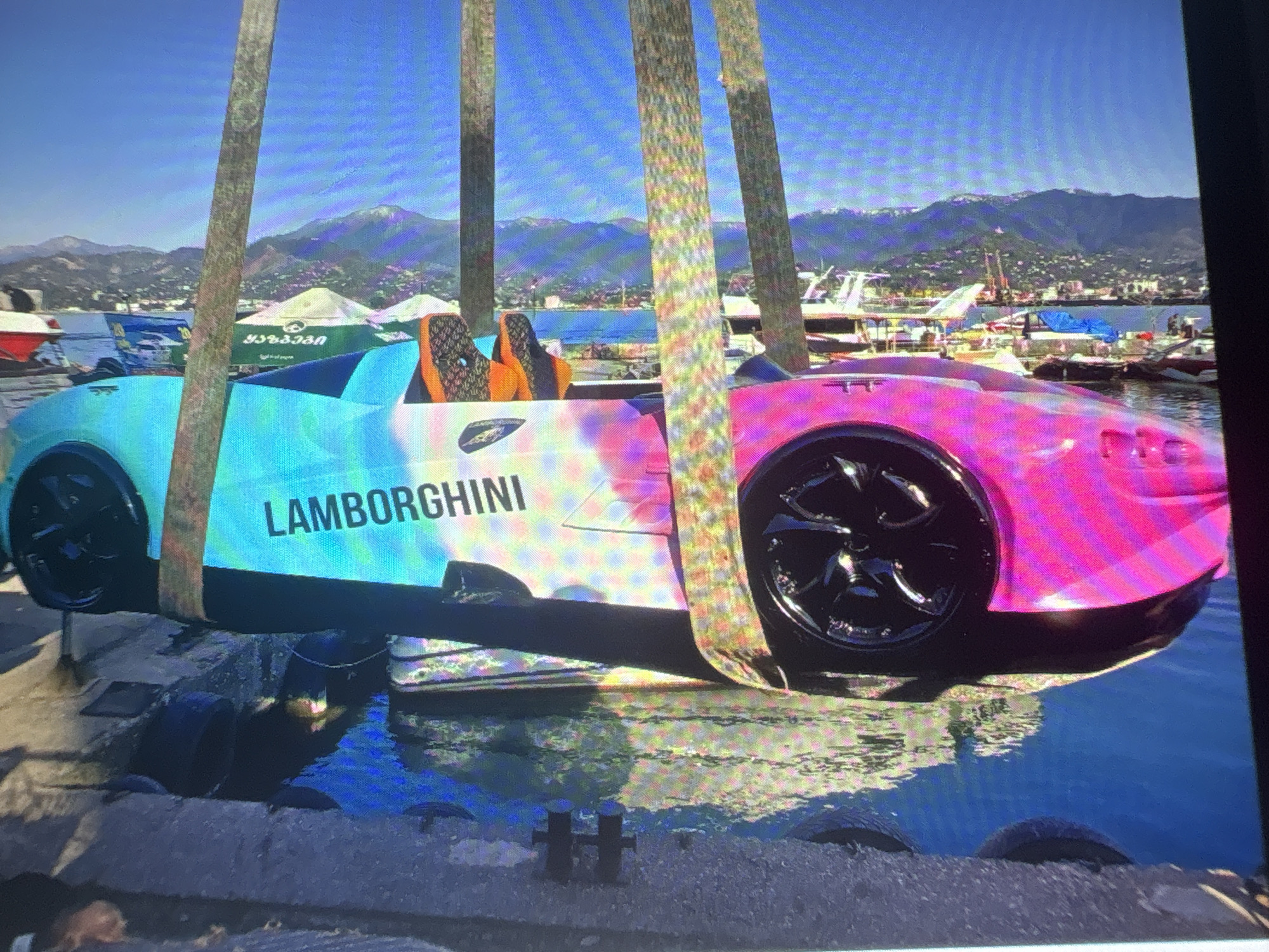 The image displays a vibrant, colorful scene that appears to be digitally rendered, possibly from a video game. In the foreground, two stylized sports cars are visible. The car closer to the viewer is a bright pink, featuring a distinctive black wheel. Behind it, another car is teal and aqua, with the word "LAMBORGHINI" printed vertically in white on its side. Both cars are positioned at an angle, suggesting they are on a road or pathway.
The background reveals a lively waterfront setting. Buildings are clustered along a coastline, and mountains rise in the distance under a clear blue sky. The overall impression is one of a sunny, bustling environment. The visual quality suggests a digital rendering, with bright colors and a stylized aesthetic. The specific location, Batumi, Georgia, is noted, which is a coastal city known for its modern architecture and Black Sea resorts.