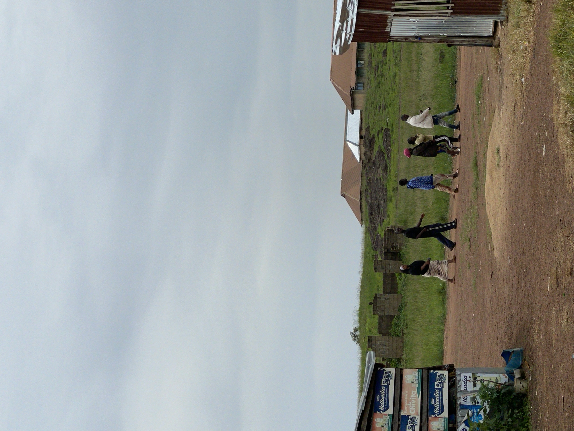 The image depicts an outdoor scene in Durumi, Nigeria, under an overcast sky. In the foreground, a dirt path runs horizontally across the frame. To the left, a makeshift stall or kiosk is visible, stacked with boxes displaying "Hollandia" branding, suggesting it sells beverages. A small plant is growing near the stall.
Further back, a group of six individuals are walking along the path. From left to right:
1. A person in a blue and white patterned shirt and light-colored pants.
2. A person in a dark jacket and patterned pants, with a pink head covering.
3. A person in a dark jacket and lighter pants.
4. A person in a white jacket and blue jeans.
5. A person in a dark top and light-colored skirt.
6. A person in a dark top and blue jeans with white stripes.
Behind the walking group, there are partially constructed brick structures and a grassy, undulating landscape. In the middle ground, several buildings with reddish-brown roofs are situated on the higher ground. To the far right, a corrugated metal structure, possibly a shelter or small building, stands prominently. The overall environment suggests a rural or semi-urban setting within Durumi, Nigeria.