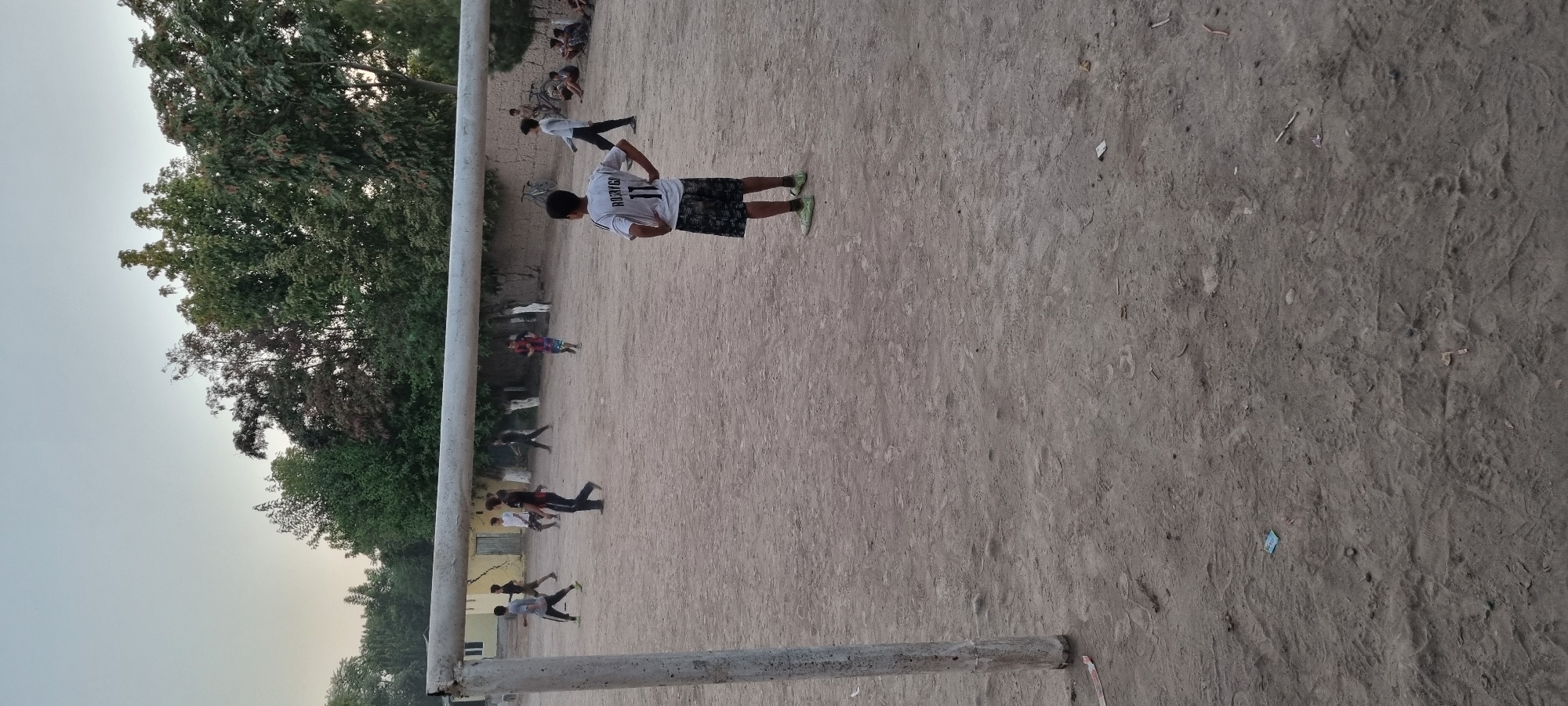 The image depicts a group of children playing soccer on a dusty field in Хаваскор, Tajikistan. The field appears to be a local park or schoolyard, with a rudimentary soccer goal constructed from metal poles. The surrounding environment includes trees and a low-lying building in the background. The sky is overcast, suggesting it is either early morning or late afternoon. The children are dressed in casual clothing and appear to be enjoying themselves. One child in the foreground is wearing a soccer jersey with the number 11.