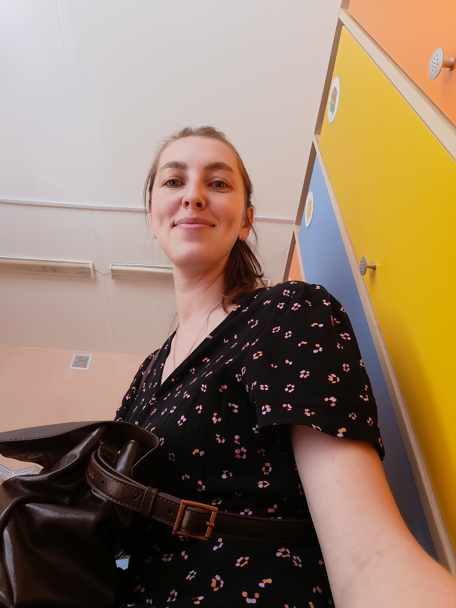 A low-angle, medium shot depicts a woman standing indoors, possibly in the city of Chaikovsky, Russia, considering the provenance of the image. The woman is fair-skinned with brown hair pulled back into a ponytail, and she is wearing a black dress with small pink and tan floral patterns. A dark brown, satchel-style bag with a buckle is visible on her left side.
The background features a set of brightly colored lockers, arranged in a vertical stack. The colors of the lockers are orange, yellow, and blue, each featuring a circular decorative sticker of an animal and a silver knob. The area above the lockers is plain white, suggesting a ceiling. The lighting is diffused. The woman is smiling slightly, looking at the camera, which gives the impression of a selfie.