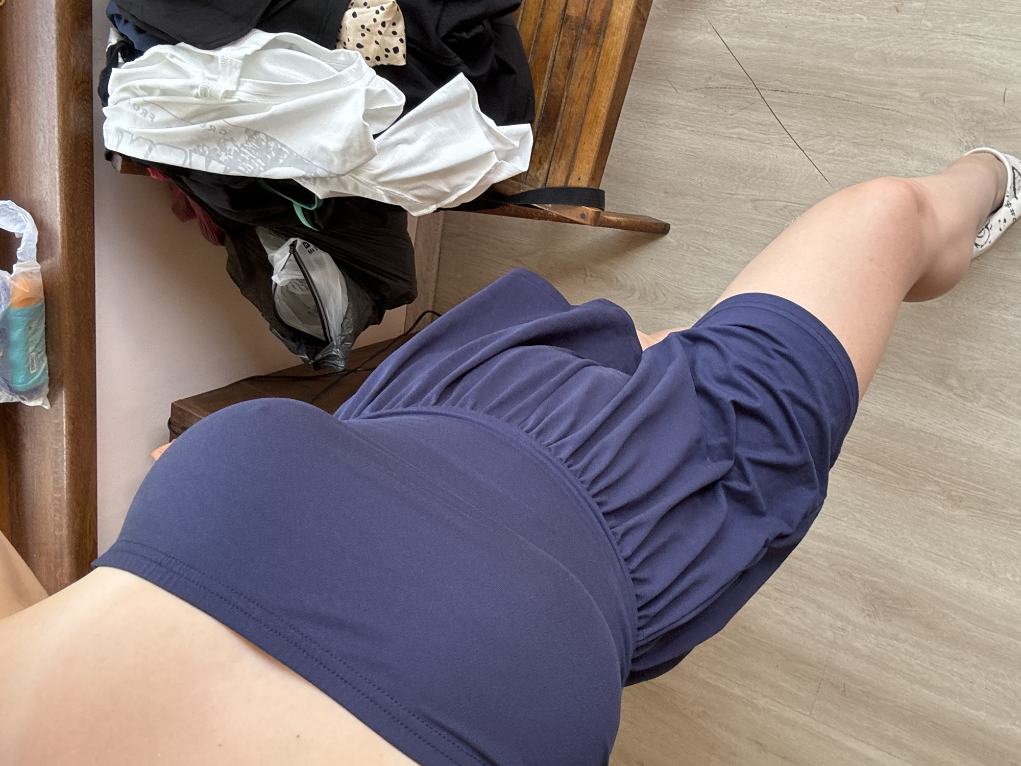 This is a high-angle, eye-level shot of a person's legs and torso, focusing on their clothing and surrounding environment. The person is wearing a navy blue ruched dress and a white slip-on shoe with a black and white pattern. Their right leg is extended forward, bent at the knee.
The background includes a wooden chair with some laundry piled on it, including a white t-shirt with a graphic print, black clothing, and a spotted fabric item. To the left of the chair, a plastic bag with an orange bottle inside is visible. The floor is made of light-colored wood planks.
The scene appears to be indoors, possibly a bedroom or dressing area. The lighting is natural, suggesting daytime. There is no discernible weather information. The overall impression is casual and domestic. The visible text on the white t-shirt is unclear due to the resolution and fold of the fabric. The image does not convey any strong emotions. The location is specified as Prodanovtsi, Bulgaria.