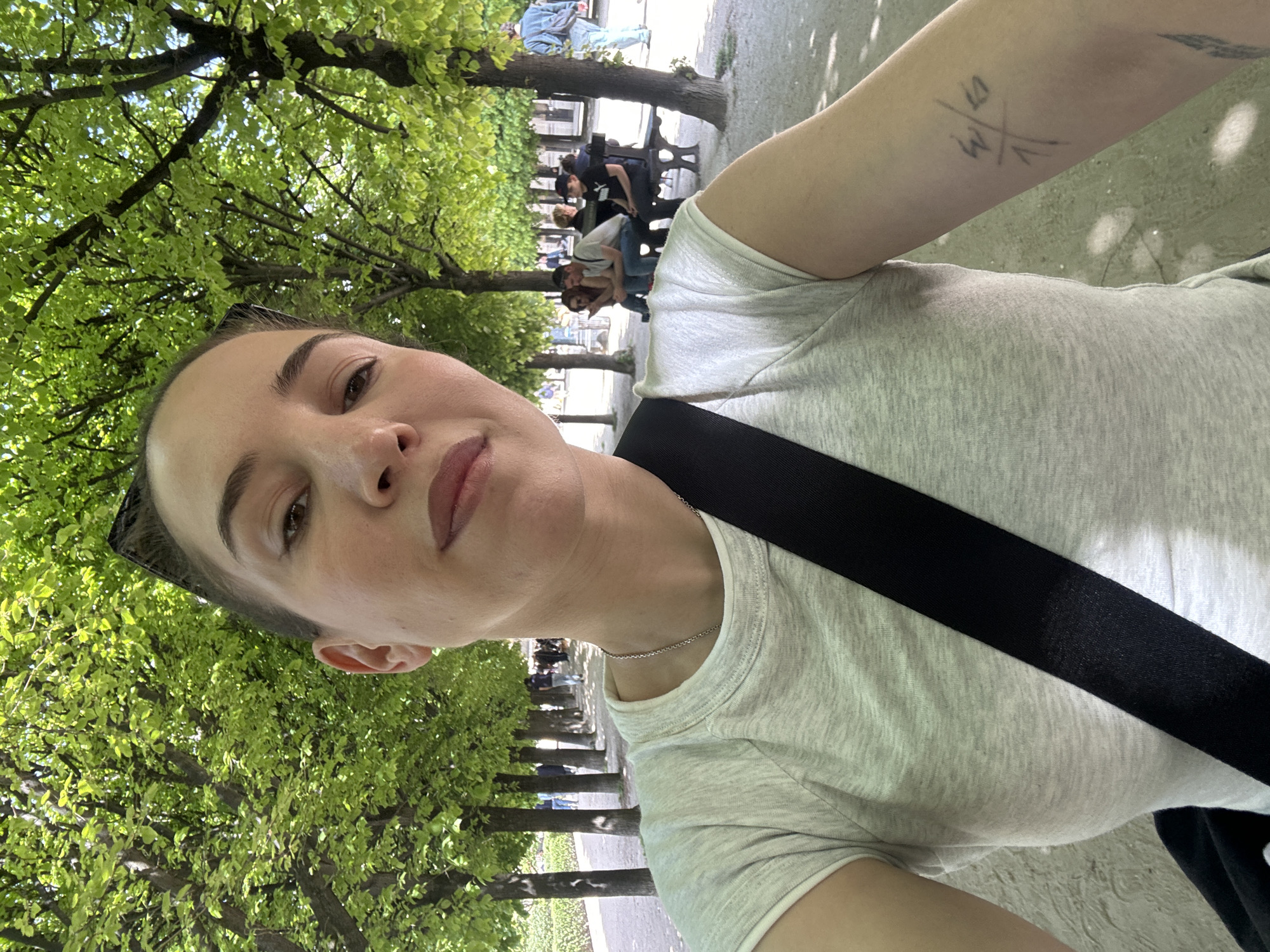 A young adult woman takes a selfie outdoors in what appears to be a park or tree-lined avenue in Paris, France. The image is taken from a slightly low, upward angle. The woman, with a neutral expression and looking directly at the camera, occupies the foreground. She has light skin, dark eyebrows, and wears a light grey t-shirt. A black strap, likely from a bag, crosses her shoulder, and a thin silver necklace is visible. Sunglasses are pushed up onto her head. On her left arm, there is a visible tattoo that appears to be a stylized "S/W" or "3/W" intersected by vertical lines.
The background features a long, straight avenue lined with mature trees boasting vibrant green foliage, indicative of spring or summer. Sunlight filters through the leaves, creating bright patches and shadows on the ground and surrounding elements. Several other people are visible in the distance, walking along the paved path or seated on dark-colored park benches under the trees. The setting suggests a pleasant, sunny daytime. The overall scene conveys a sense of leisurely activity in a well-maintained urban green space.