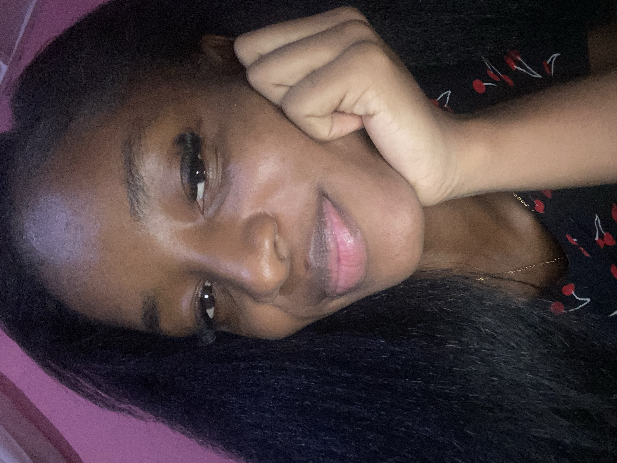 A close-up, head-and-shoulders selfie of a young Black woman is presented. She has dark, long hair that frames her face, and her left hand is resting on her chin. She is looking towards the camera with a slight smile. Her clothing is visible in the lower right corner, a dark fabric with a cherry pattern. The background is a soft pink, suggestive of a wall or fabric. The lighting is warm and appears to be artificial, casting a slight sheen on her skin. The image is taken indoors, likely in a residential setting. There is no specific information within the image to indicate its location in Jos, Nigeria.