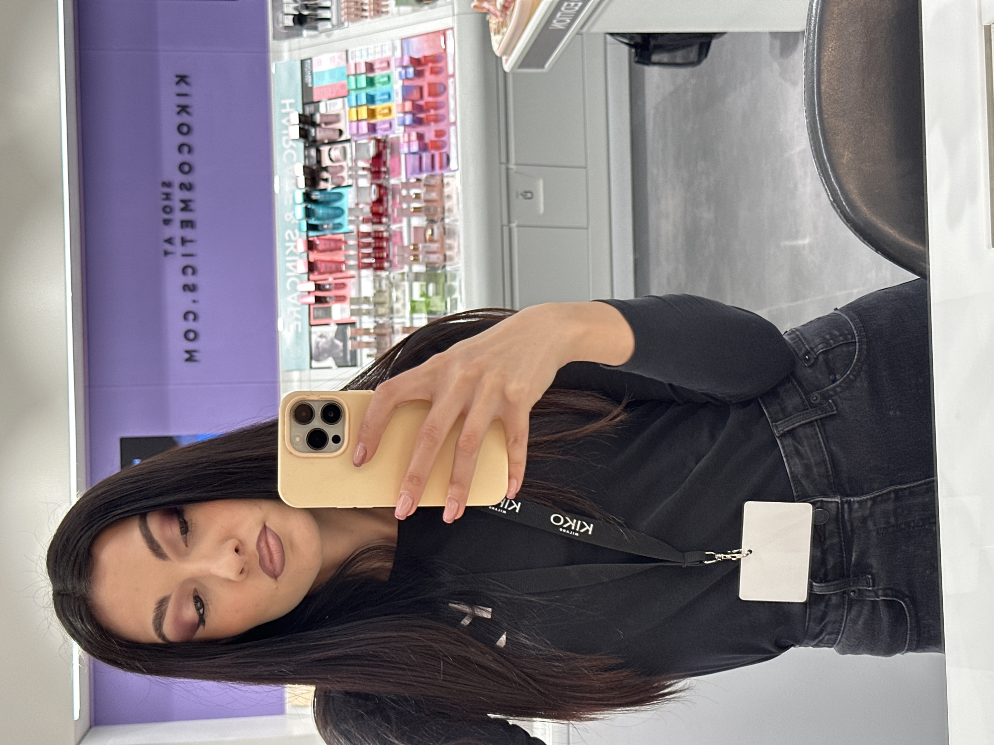 An eye-level mirror selfie captures a young woman with long, dark hair in a cosmetics store. She is holding a light yellow smartphone, likely an iPhone, in her right hand to take the picture. She is wearing a black long-sleeved top and dark jeans. A black lanyard with "KIKO" written on it hangs around her neck, attached to a blank white ID card or badge. Her makeup features smokey purple eyeshadow and nude-toned lipstick, and her nails are painted a light pink.
The background, visible as a reflection, shows a brightly lit retail environment. On the left, a purple wall displays mirrored text that reads "KIKOCOSMETICS.COM" and "SHOP YA" (reversed from "AY POHS"). Beyond this, shelves are stocked with various colorful makeup and skincare products. The floor is a light grey, and other commercial fixtures are partially visible. The overall scene suggests an indoor setting, likely a KIKO Cosmetics store. The image is oriented horizontally, rotated 90 degrees counter-clockwise from a standard portrait view.