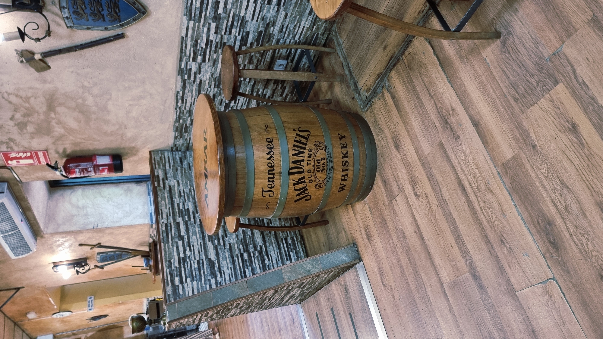 The image depicts an indoor scene, likely a bar or restaurant, with a distinct rustic aesthetic. The focal point is a round table made from a wooden barrel, branded with "Jack Daniel's Tennessee Whiskey" and "Ambar". Two wooden stools are positioned around the table. In the background, a wall is decorated with a stone-like tiled pattern and textured plaster. A red fire extinguisher is mounted on the wall, along with decorative items such as a shield and an axe. The flooring is made of wooden planks. The overall impression is one of a cozy, themed establishment. There are no people visible in the image. The location in the city of Besalú, Spain, is not evident from the visual information provided.