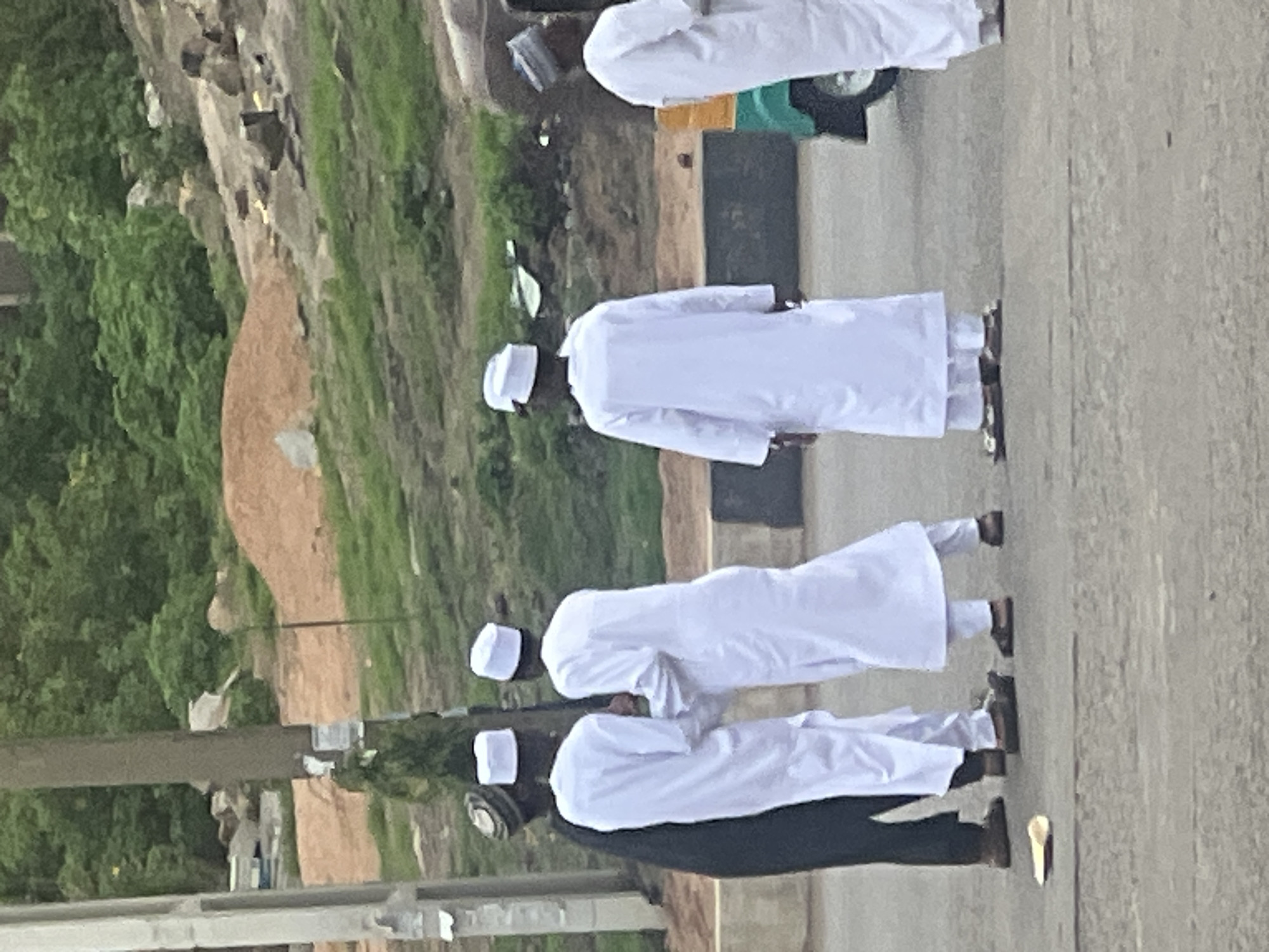 Four people are walking on a paved street in Gura Top, Nigeria. They are wearing white robes and white caps, except for one person in the front who is wearing a black robe. The surrounding area includes a hill with grass and trees. A vehicle is parked on the side of the road.
