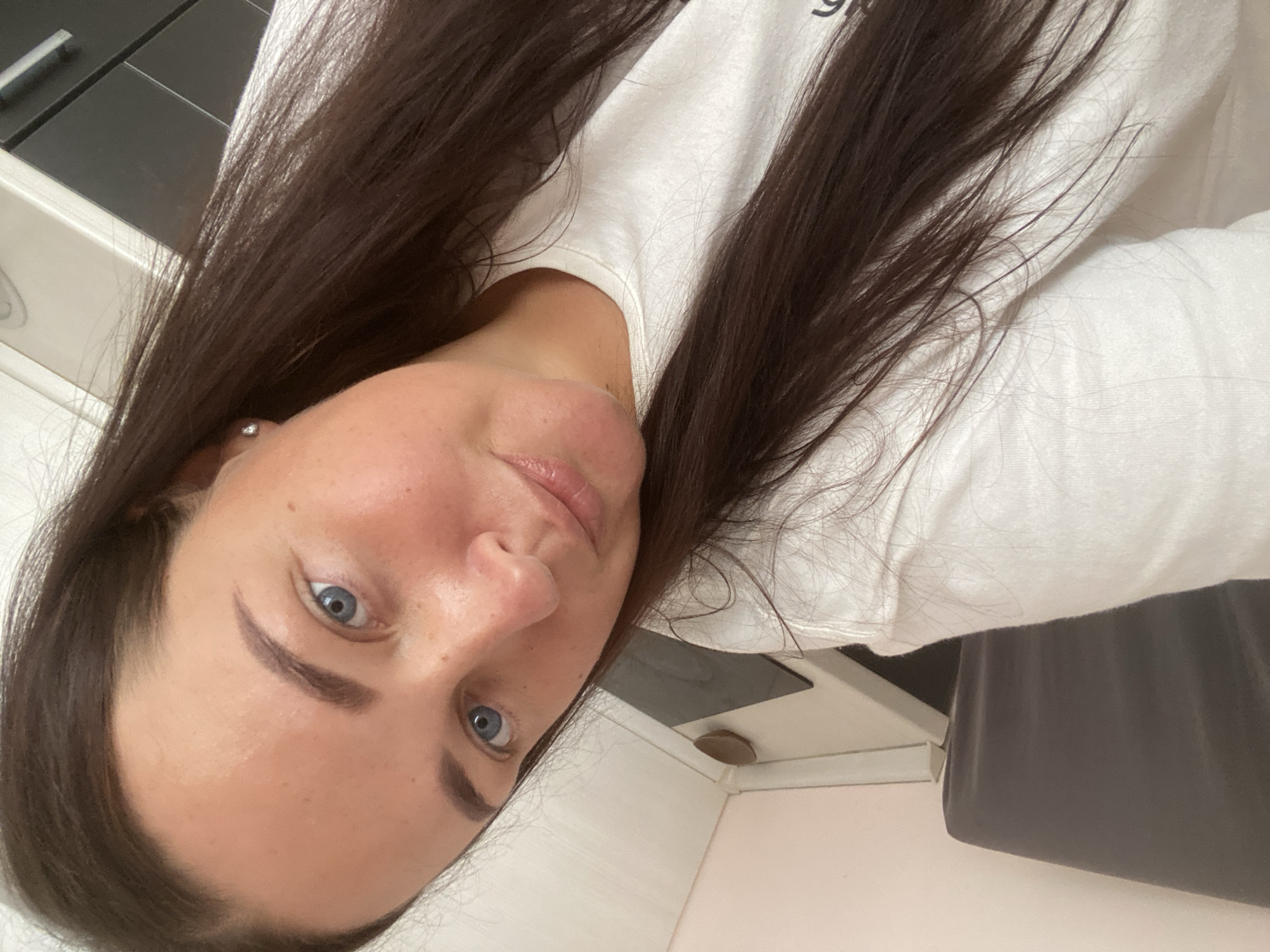 A young woman with long, dark brown hair and blue eyes is captured in a close-up, slightly tilted selfie. She is wearing a plain white t-shirt. The background appears to be an indoor setting, possibly a kitchen or bathroom, with hints of cabinetry and a pale wall. The lighting suggests an ambient indoor environment, and the focus is entirely on the woman's face and upper torso. No specific location in Sochi, Russia, is identifiable from the provided image.