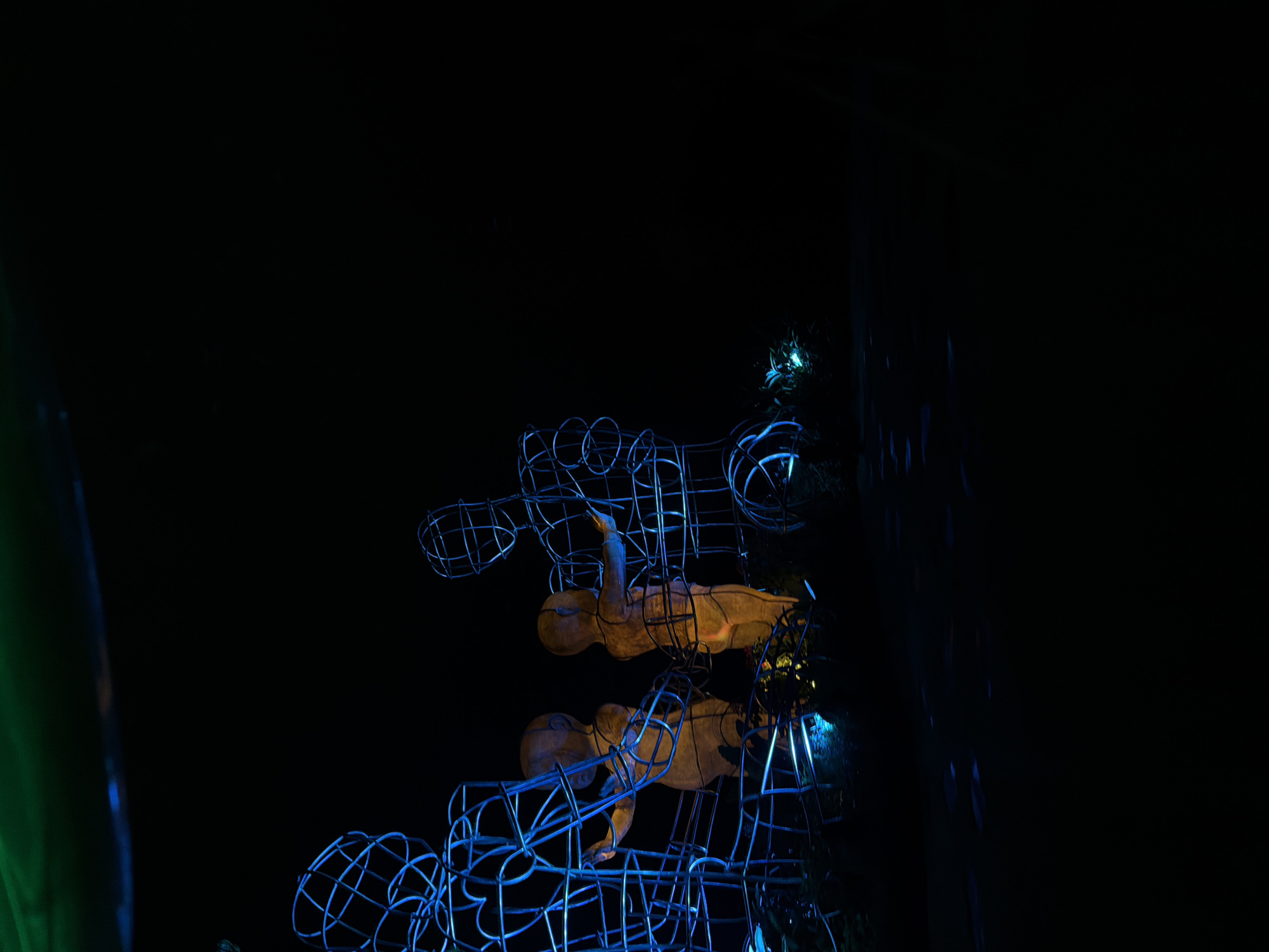The scene depicts an outdoor art installation at night in the city of Unknown, Indonesia. Centrally positioned are two solid, child-like figures illuminated with a warm amber glow. Each child figure extends an arm towards an adjacent larger, upright human-shaped sculpture constructed from blue-illuminated wireframe. A wireframe figure stands to the left of the left child, and another to the right of the right child. The ground beneath the sculptures is dark, marked by scattered small blue lights and faint outlines of vegetation. The background is predominantly black, with a subtle green light visible in the upper left quadrant. Artificial lighting highlights the sculptures against an otherwise dark environment.