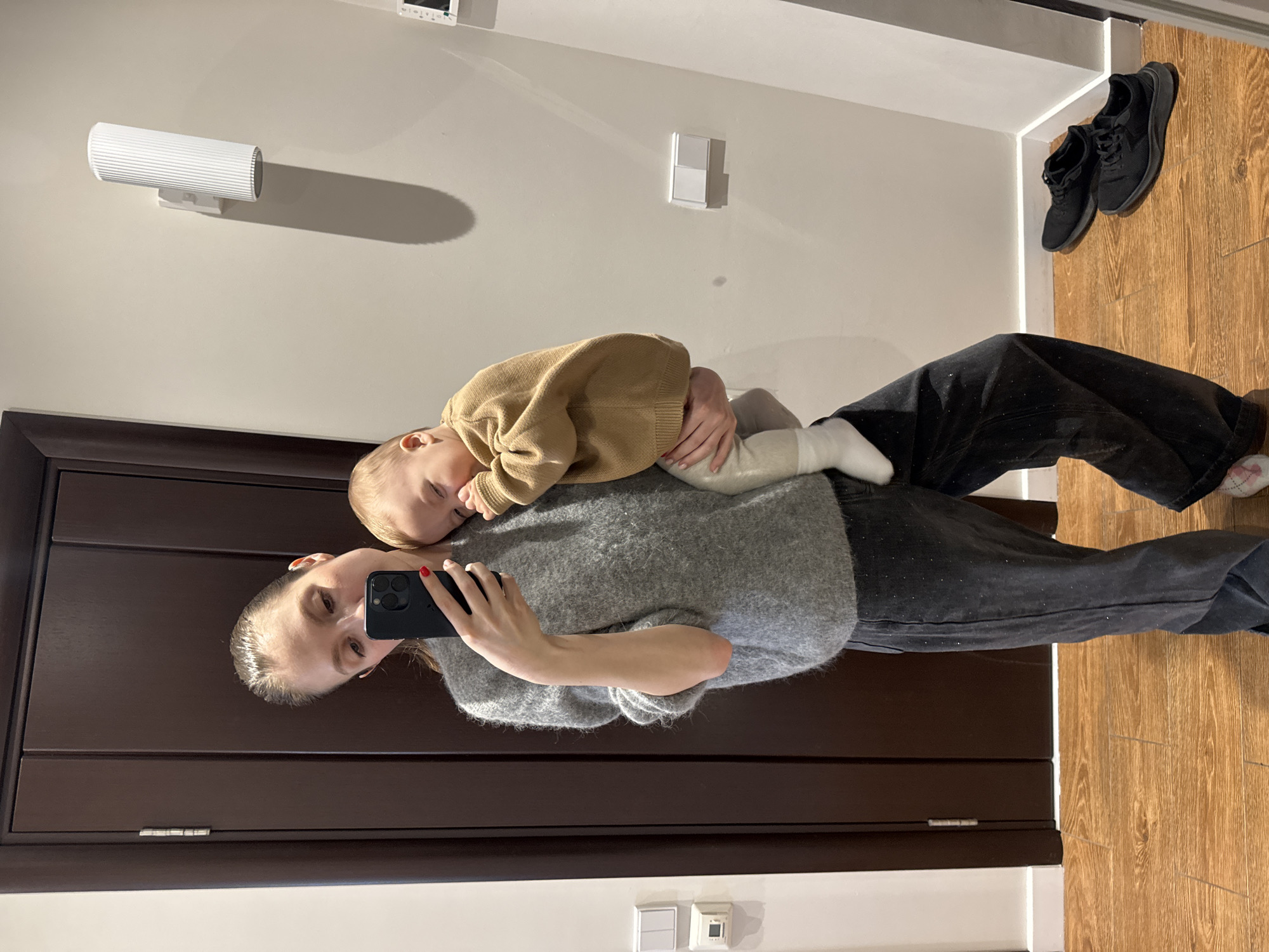 A woman holds a baby in her arms, appearing to take a selfie in front of a mirror. The woman has light skin and fair hair. She is wearing a grey sweater and dark pants. The baby, also with light skin, wears a brown sweater and light-colored pants and socks. The room features a dark-colored door behind the woman, a white wall with light fixtures and electrical outlets, and a wooden floor with black shoes placed against the wall. The setting appears to be inside a home. There is no definitive evidence that this image was taken in Moscow, Russia.