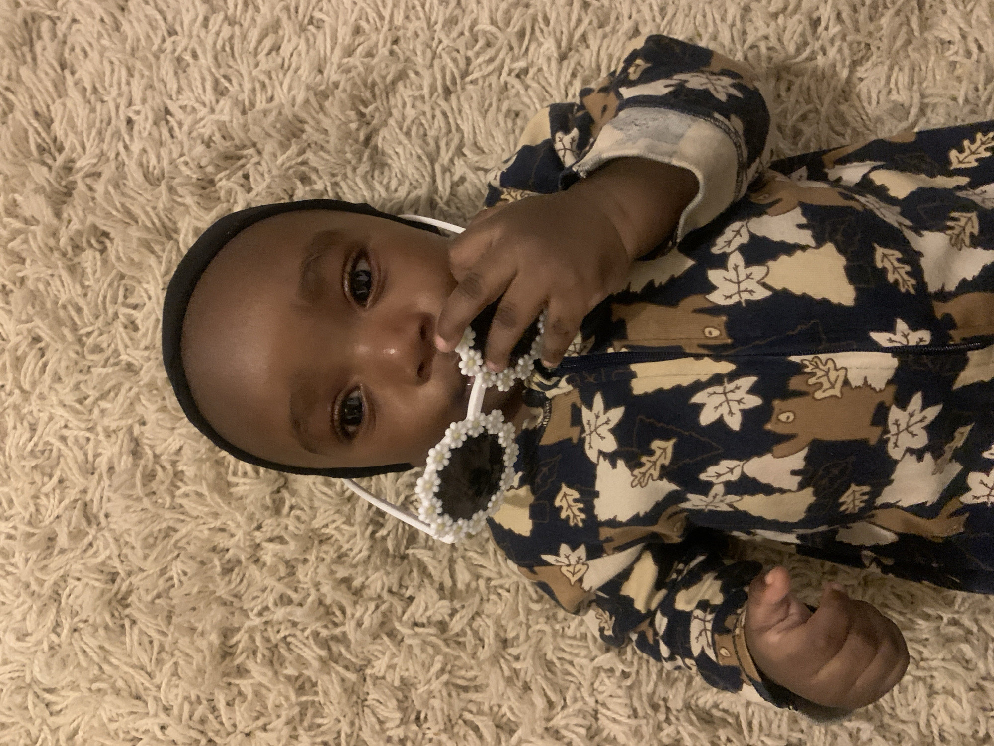 A baby lies on a light-beige carpet wearing a black headband and a navy-blue and beige patterned onesie. The baby holds a pair of white sunglasses with flower-shaped details on the frames in their hand, near their mouth. The location and city in Nigeria are not discernible from this image.