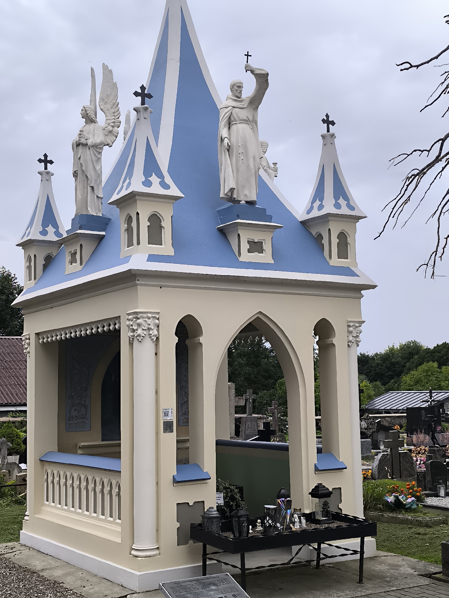 A beige and light blue ornate mausoleum with a pointed roof stands in a cemetery in Kretinga, Lithuania. The mausoleum is decorated with white Gothic-style architectural elements, including finials, arches, and columns. Statues of an angel and a robed figure holding a cross adorn the roofline. In front of the mausoleum, a black metal table holds various memorial items, including lanterns, candles, and small figurines. The background shows rows of gravestones and trees, typical of a cemetery setting. The sky is overcast.