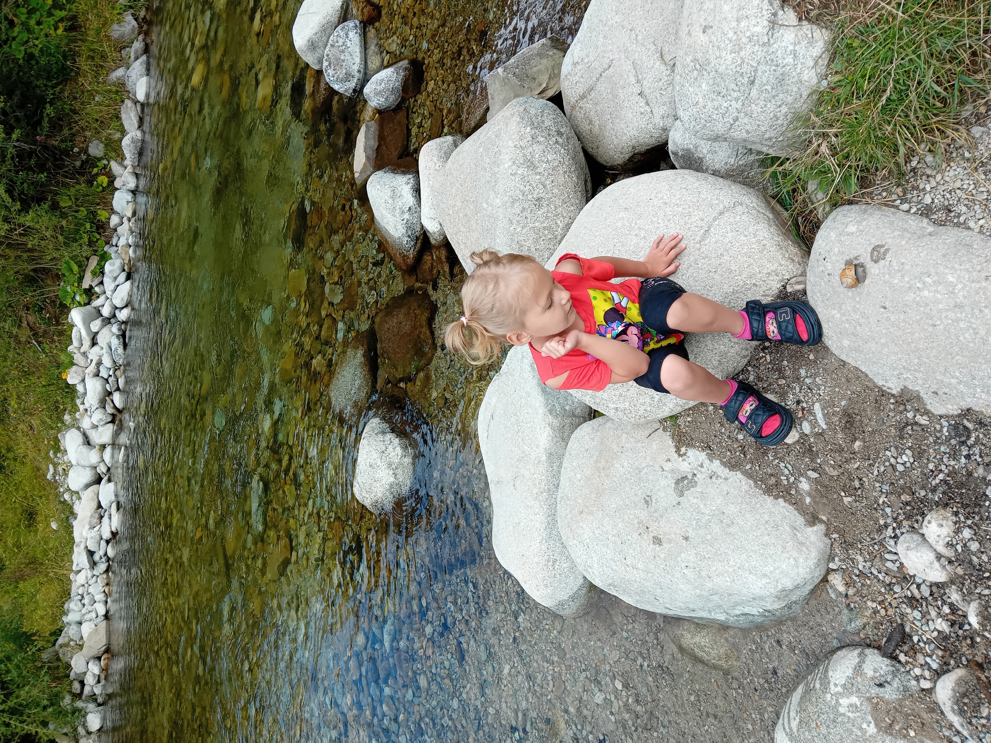 A young girl with blonde hair in two pigtails sits on large, smooth river stones near the bank of a clear, shallow river in Zuberec, Slovakia. She is wearing a red off-the-shoulder t-shirt with a graphic print and black shorts, as well as black sandals with pink socks. Her right hand is resting on her chin as she looks down. The riverbed is visible through the transparent water, showing a mix of small pebbles and larger stones. The riverbank is lined with more smooth, rounded rocks, and beyond that, lush green grass and trees. The overall scene depicts a serene natural setting.