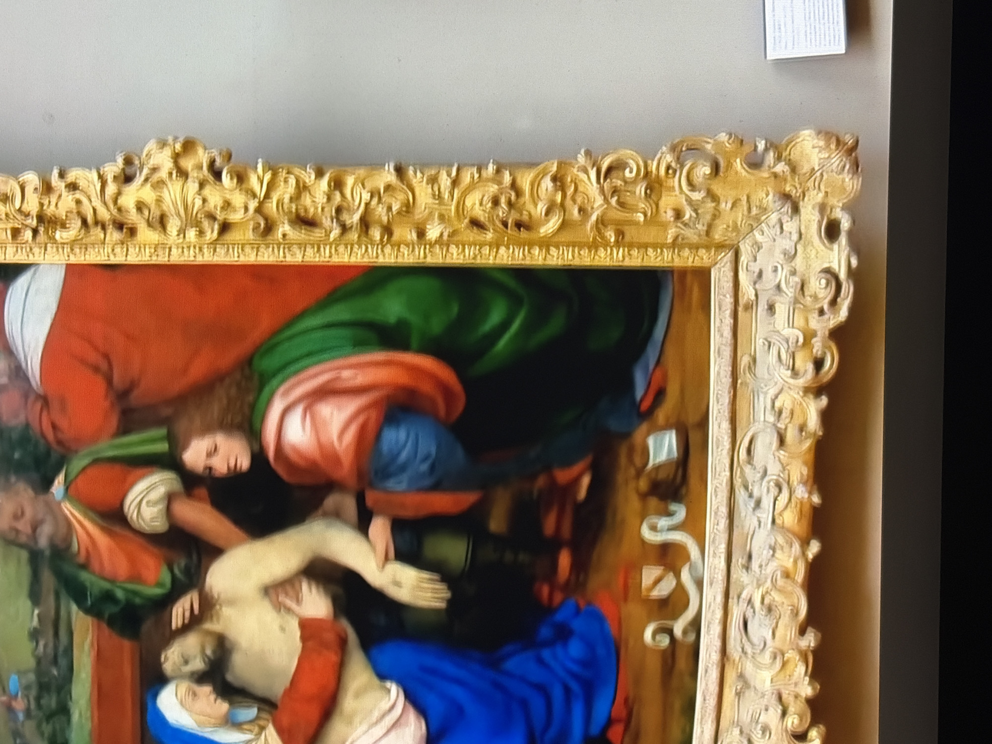 An ornate gold frame surrounds a detailed painting depicting a religious scene. In the foreground, a pale-skinned man, presumably Jesus Christ, lies in the arms of a woman wearing a blue garment and a white head covering, likely the Virgin Mary. Another woman in a green robe and a man in a red robe with a green overlay stand behind them, observing the scene. The painting features rich colors and intricate details in the figures' clothing and expressions, suggesting a period artwork. The frame itself is elaborate, with carved scrollwork and decorative elements. In the lower right corner, a small white information placard is partially visible, indicating the artwork's presence in a public space, such as a museum.
