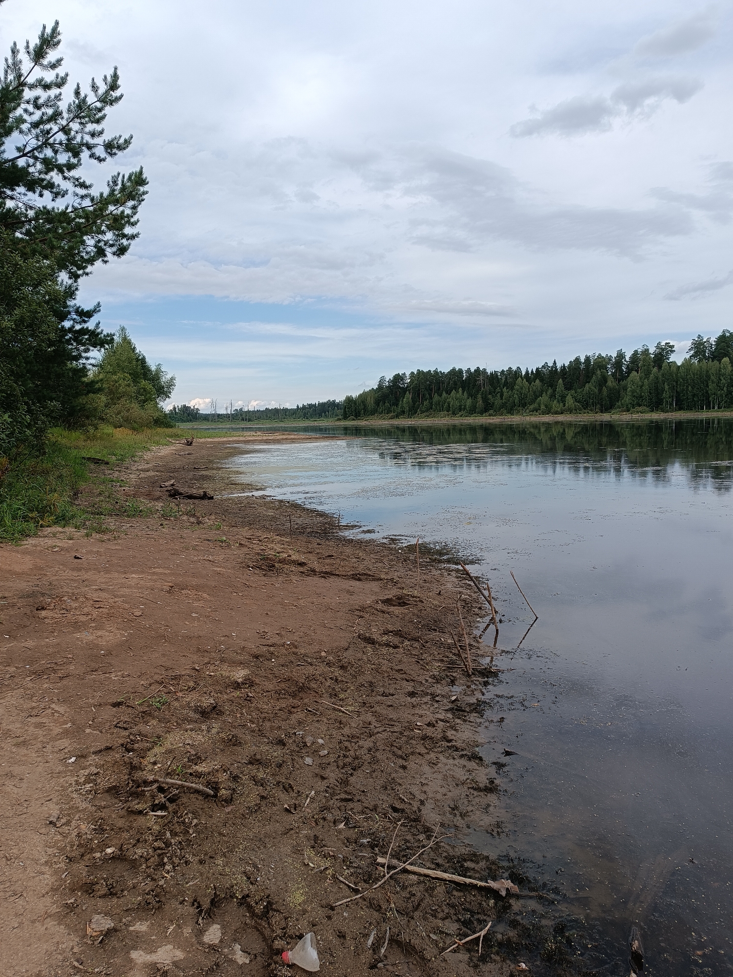 A wide, calm lake is shown in the foreground with a dirt bank on the left. The bank is covered in dry earth, scattered debris, and some sparse vegetation. Beyond the lake, a dense forest lines the horizon under a cloudy sky. The scene is set in Opary, Russia, suggesting a natural, possibly remote, location. There are no people visible in the image.
