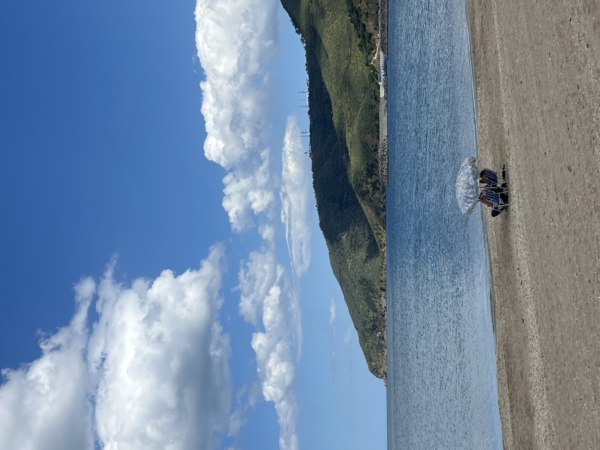 The image presents a coastal scene, likely in the city of M'Diq, Morocco. The view encompasses a beach with a sandy shore meeting the blue water. People are sitting in chairs under an umbrella on the beach. A green mountain range rises in the background, partially obscuring the sky filled with fluffy white clouds. The sky has varying shades of blue. Overall, the scene evokes a sunny and tranquil beach day.