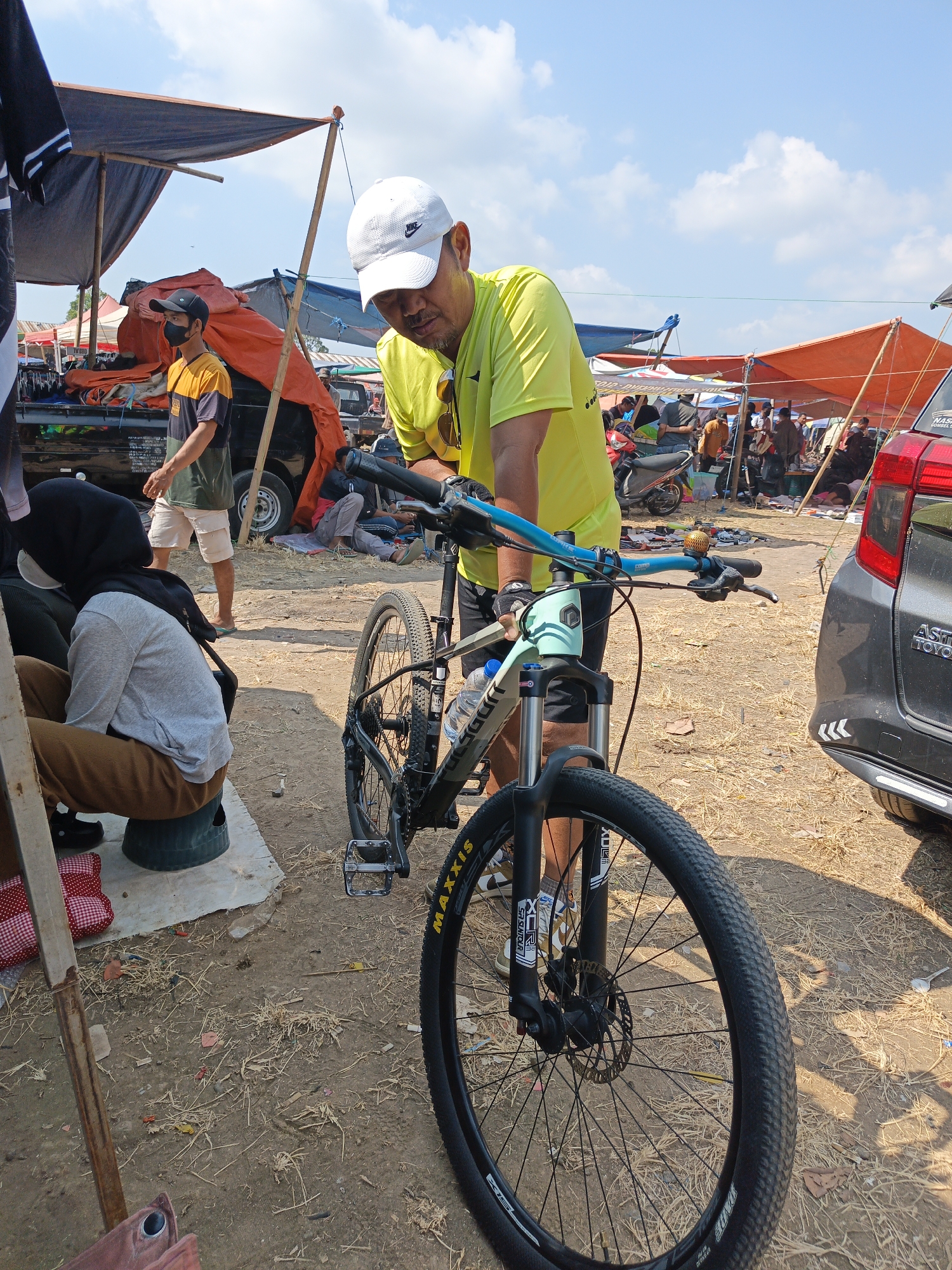In Jatinom, Indonesia, an outdoor market scene unfolds under a bright, cloudy sky. A man wearing a yellow t-shirt and a white baseball cap is leaning over a light blue mountain bike, appearing to adjust or examine it. The bike has a black frame with light blue accents and a "MAXIS" tire. In the background, various stalls are set up with orange and blue tarpaulins providing shade. People are milling about, and several motorcycles are visible. To the right, the rear of a dark grey Toyota car is parked. On the left side of the image, a person wearing a black hijab and a grey sweater sits on a green stool, with another person standing behind them. The ground is covered in dry grass and dirt, indicative of a rural or semi-rural market setting.