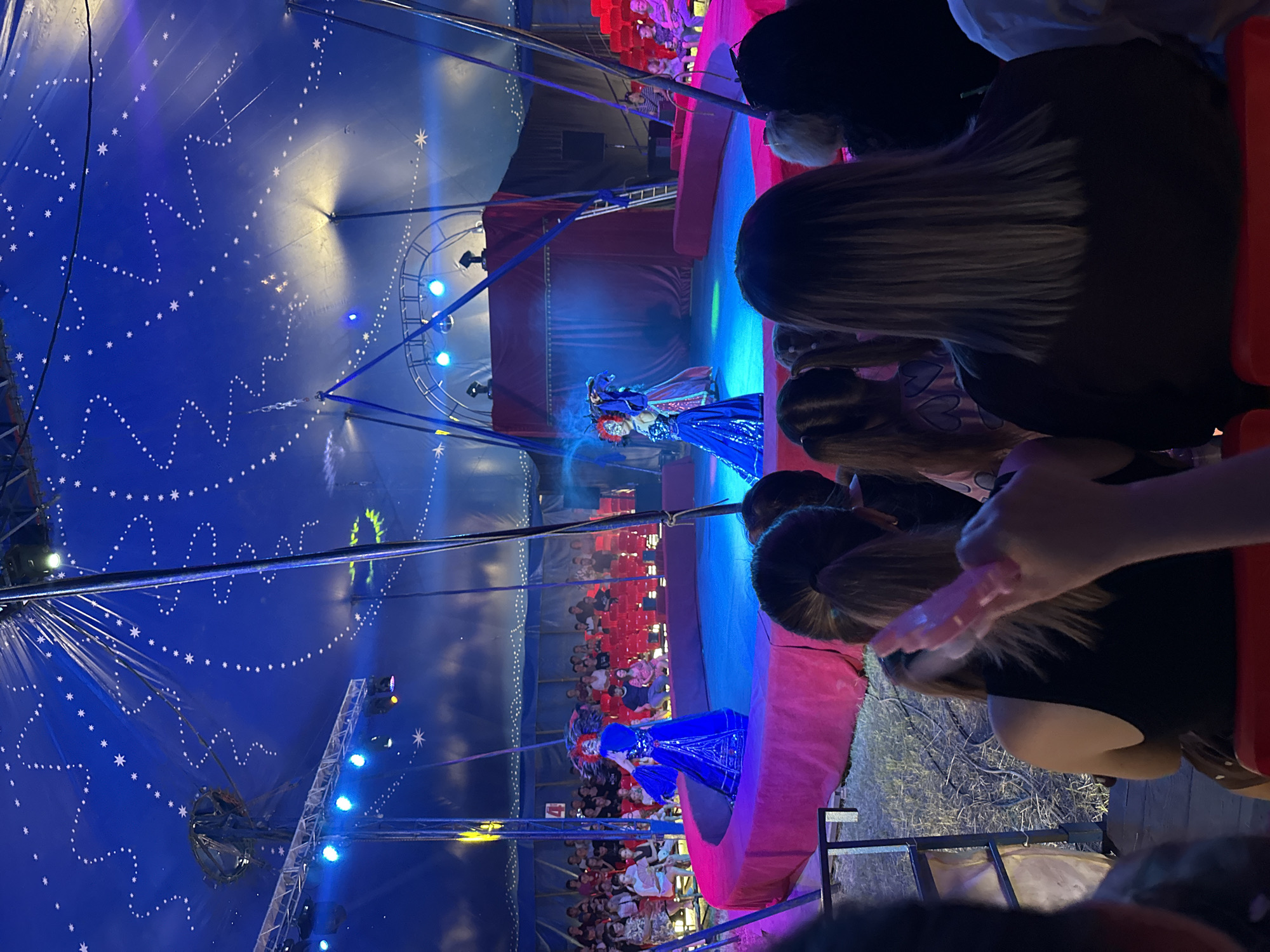 In Novoaltaysk, Russia, a circus tent is set up for a performance. The tent's interior is illuminated by a starry blue canopy and stage lights. In the center of the tent, a circular stage is adorned with a pink border. Two performers in vibrant blue and red costumes are on stage, one holding a feathered headdress. Behind them, a red curtain hangs, framing the performance area. The audience, seated in red chairs, watches the show. Spectators in the foreground are partially visible, with one person holding a pink object. The overall atmosphere is one of entertainment and shared experience under the circus tent.