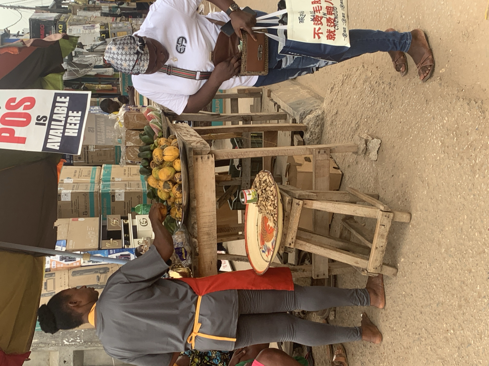 This image captures a lively outdoor market scene in Shagamu, Nigeria, under bright daylight conditions. The ground is unpaved and dusty, characteristic of a bustling street vendor area.
Two individuals are visible. In the foreground, a vendor, seen from the back/side, wears a grey shirt, grey leggings, and a red apron tied with a yellow string, standing barefoot behind a wooden market stall. The stall is laden with fresh produce, including a pile of yellow fruits, likely papayas, and green vegetables, possibly cucumbers. Adjacent to the produce, a round, decorative metal tray holds a mound of peanuts in their shells, with a small can nestled among them.
Facing the stall, presumably as a customer, is another person dressed in a white t-shirt, blue jeans, and brown sandals, wearing a patterned headscarf and a side bag with a green and red striped strap. This individual has a gold-colored watch and is reaching into a brown wallet or purse, while also holding a white reusable shopping bag. The bag prominently features Chinese characters, including "張动", "不變毛肚", "就烫鴨人", and the city name "CHENGDU".
In the background, numerous cardboard boxes are stacked high, suggesting a storage or wholesale area. Some boxes display partial English text such as "MEC", "AQ", "ens", "G", "C", and various numbers. A white and black sign on the left clearly announces "POS IS AVAILABLE HERE", indicating that Point of Sale services are offered at this location. The overall atmosphere appears routine and transactional, with no strong emotions discernible from the subjects.