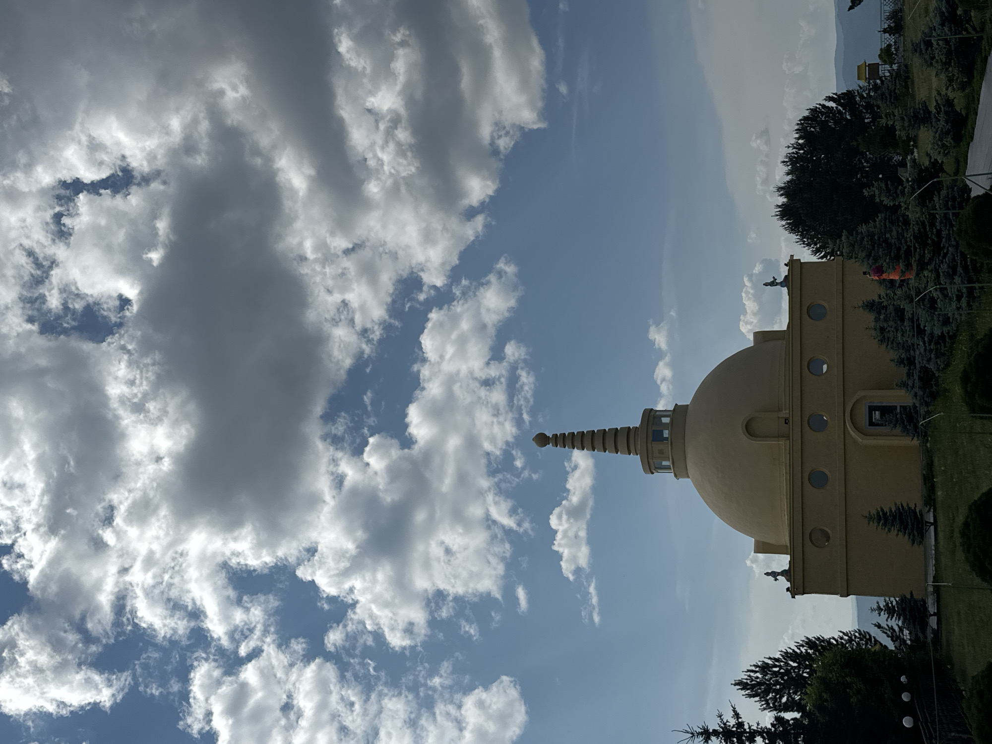 The image shows a Buddhist stupa, likely the Gandan Tegchinlen Monastery in Ulan-Ude, Russia, against a backdrop of a cloudy blue sky. The stupa is a prominent yellow dome structure with a tiered spire. There are small statues on the roof of the building section of the stupa. In the foreground, there are various coniferous trees and lush greenery. A pathway is visible to the right, and in the distance, rolling hills and a hazy landscape can be seen. A person wearing an orange garment is visible amongst the trees on the right side of the stupa.