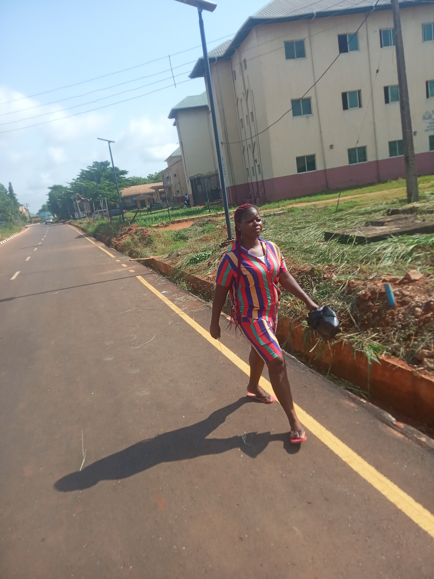 The image depicts a woman walking along the side of a paved road under bright sunshine in Nsugbe, Nigeria. The woman, positioned towards the right of the frame, is walking from left to right. She is wearing a vibrant, vertically striped two-piece outfit featuring a short-sleeved collared shirt and matching shorts in multiple colors including red, blue, green, and yellow. Her hair is braided and appears reddish-brown, and she wears red flip-flops. In her right hand, she carries a dark, indeterminate object that could be a small bag or helmet.
The road features a continuous yellow line separating the shoulder from the main lane, and a broken white line further in the distance. To the right of the woman and the road is a rough, grassy, and dirt verge with a concrete drainage channel. Beyond this, several multi-story buildings with light-colored walls and dark roofs are visible, interspersed with green trees and vegetation. The clear sky is traversed by power lines and lined with tall streetlights.
The scene suggests a sunny day, likely mid-morning or late afternoon, indicated by the long shadows cast by the woman and other objects. The environment appears to be an outdoor, possibly campus or semi-urban setting. No discernible text is visible on the buildings or elsewhere in the image.