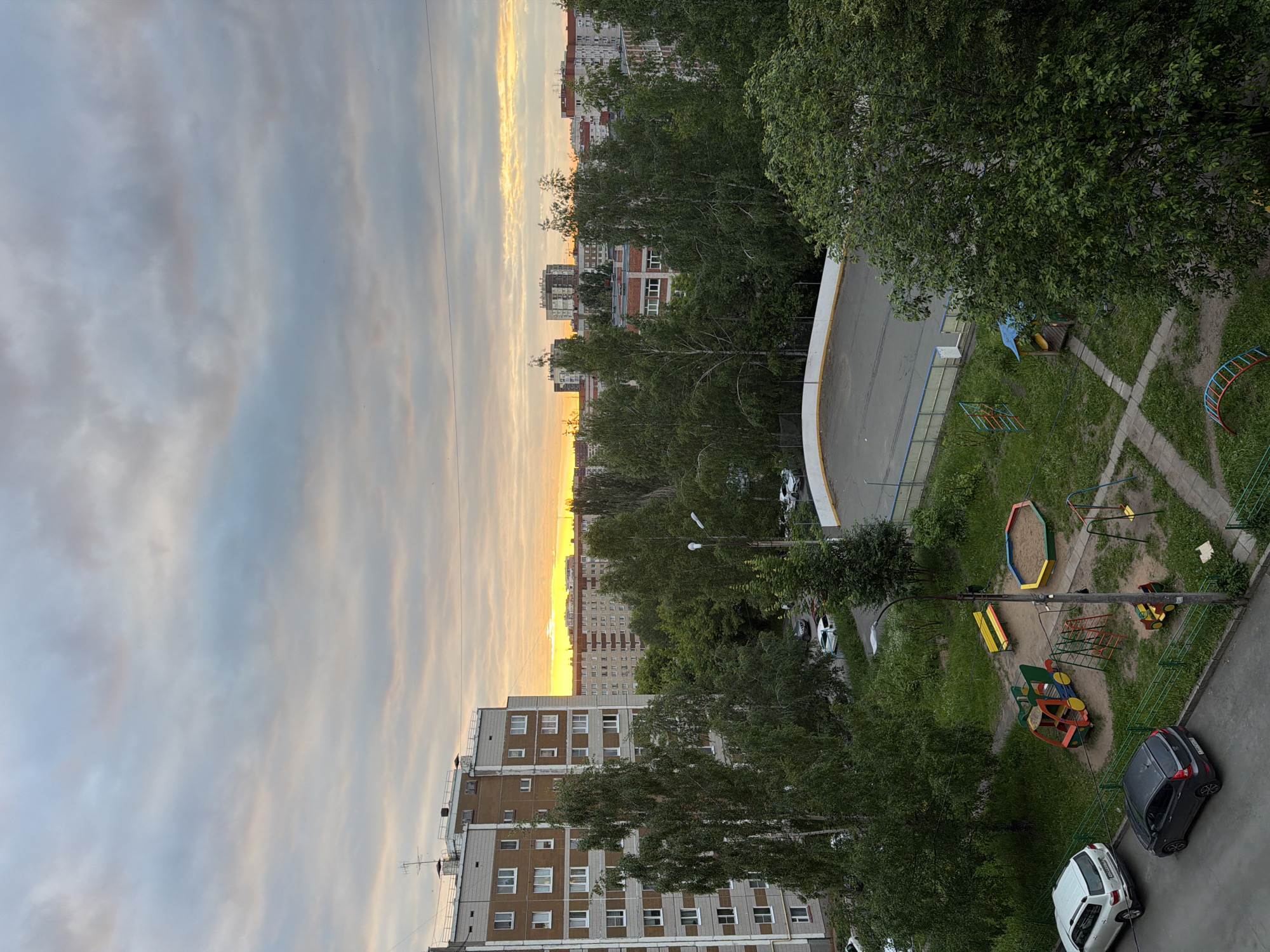 An outdoor daytime scene in a residential area of Izhevsk, Russia, depicts a view from a building overlooking a courtyard. The sky is partly cloudy with a golden sunset visible on the horizon. In the foreground, a children's playground area features a sandbox, a multi-colored play structure resembling a vehicle, swings, and a small yellow and red toy car. Adjacent to the playground is a paved area, possibly a sports court, with a white border and blue railing. Several trees with lush green foliage are scattered throughout the scene. Parked cars, including a white SUV and a dark hatchback, are visible on a paved surface. In the background, several multi-story apartment buildings are present, consistent with the urban landscape of Izhevsk.
