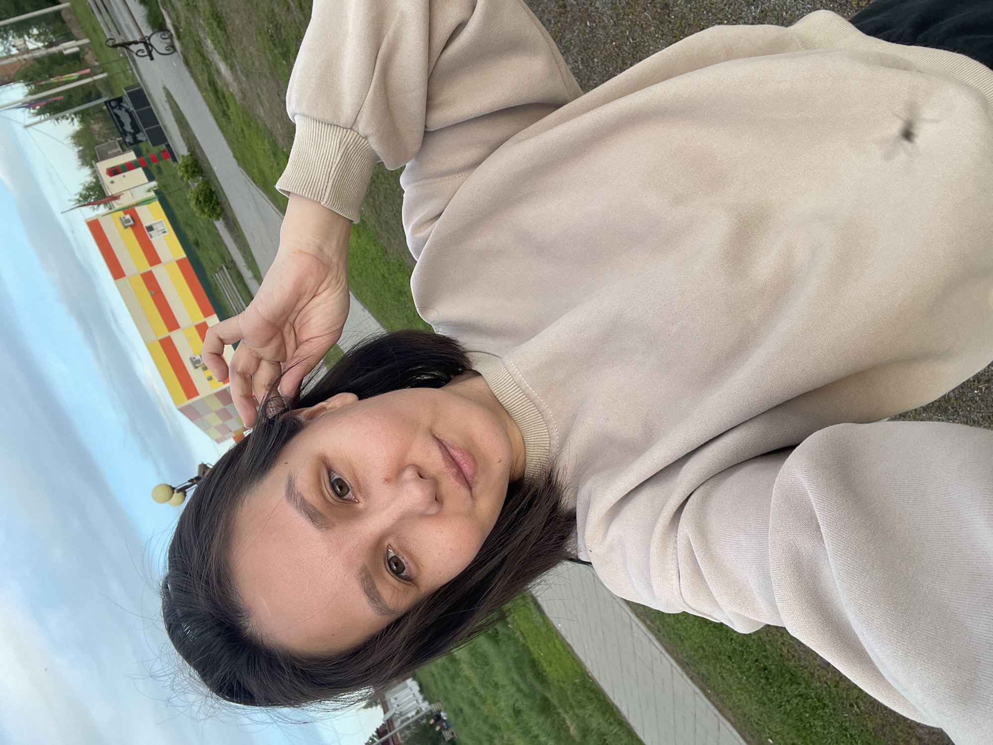 A woman is taking a selfie outdoors. She is wearing a beige sweatshirt and has dark hair. In the background, there is a tall, narrow building with yellow, red, and white stripes, indicating the location is Verkh-Katunskoye, Russia. There is also a paved path, green grass, and a street lamp visible. The sky is cloudy.