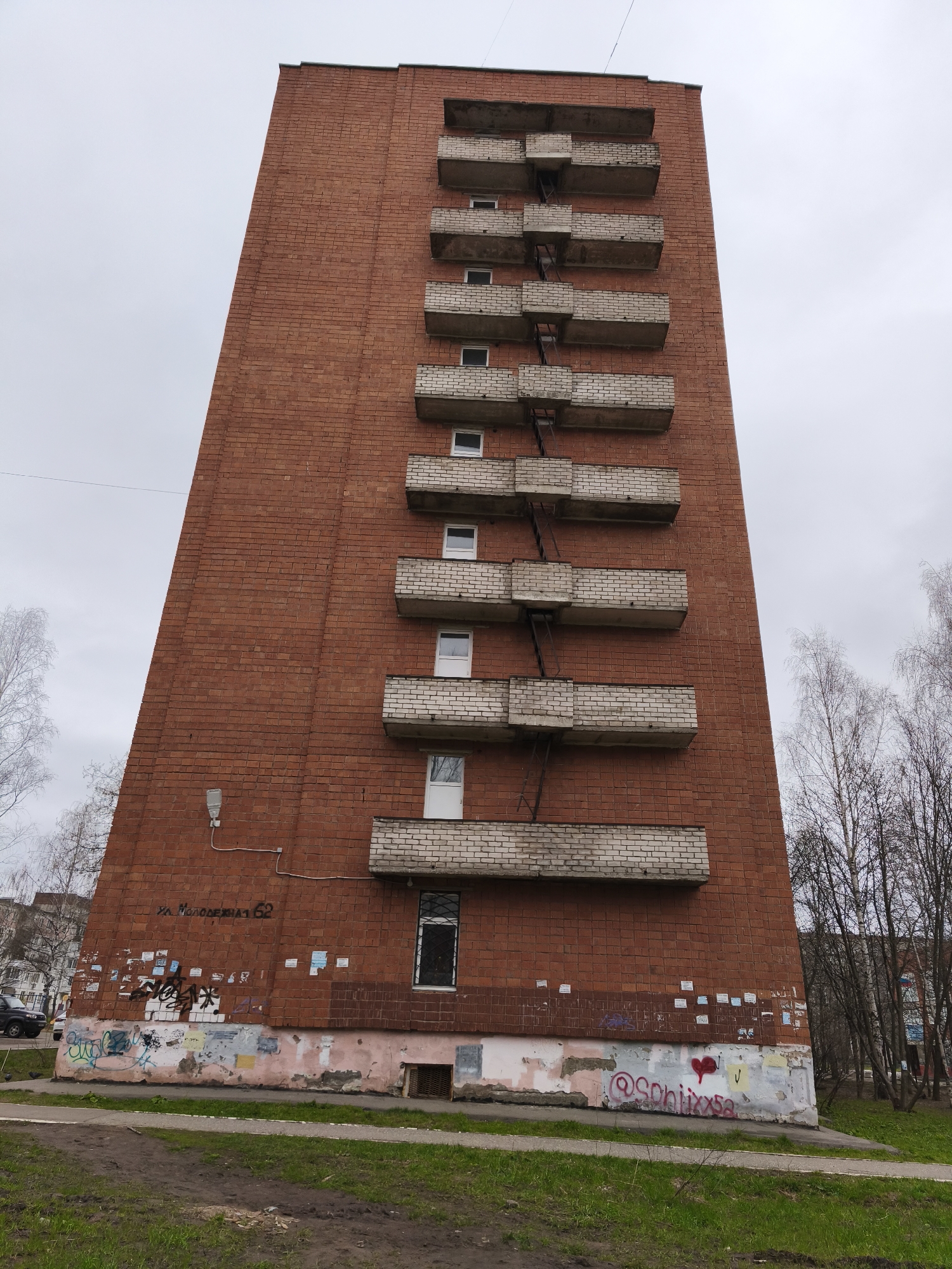 The image displays a tall, narrow residential building constructed from reddish-brown brick, set against an overcast, grey sky. The building features nine visible floors, each with a small, white-framed window and an attached balcony with light-colored brick railings. A black, ladder-like structure, likely a fire escape or maintenance access, zig-zags down the side, connecting the balconies.
No specific activity or event is taking place; the image is a static depiction of the building and its immediate surroundings.
Notable details include the visible signs of wear and age on the building, especially its base, which shows peeling paint, old advertisements, and extensive graffiti. A single street lamp is mounted on the brick wall to the left of the balconies. The bare branches of deciduous trees, devoid of leaves, flank the building, suggesting a cold season such as late autumn, winter, or early spring. The overall architectural style and condition are typical of older residential blocks in post-Soviet urban environments. The location is Izhevsk, Russia.
Visible text on the lower section of the brick wall includes "ул. Молодежная 62" written in Cyrillic. Below this, amidst the graffiti, is "@Schixxsa" accompanied by a red heart symbol. Other less legible graffiti tags are also present on the painted base.