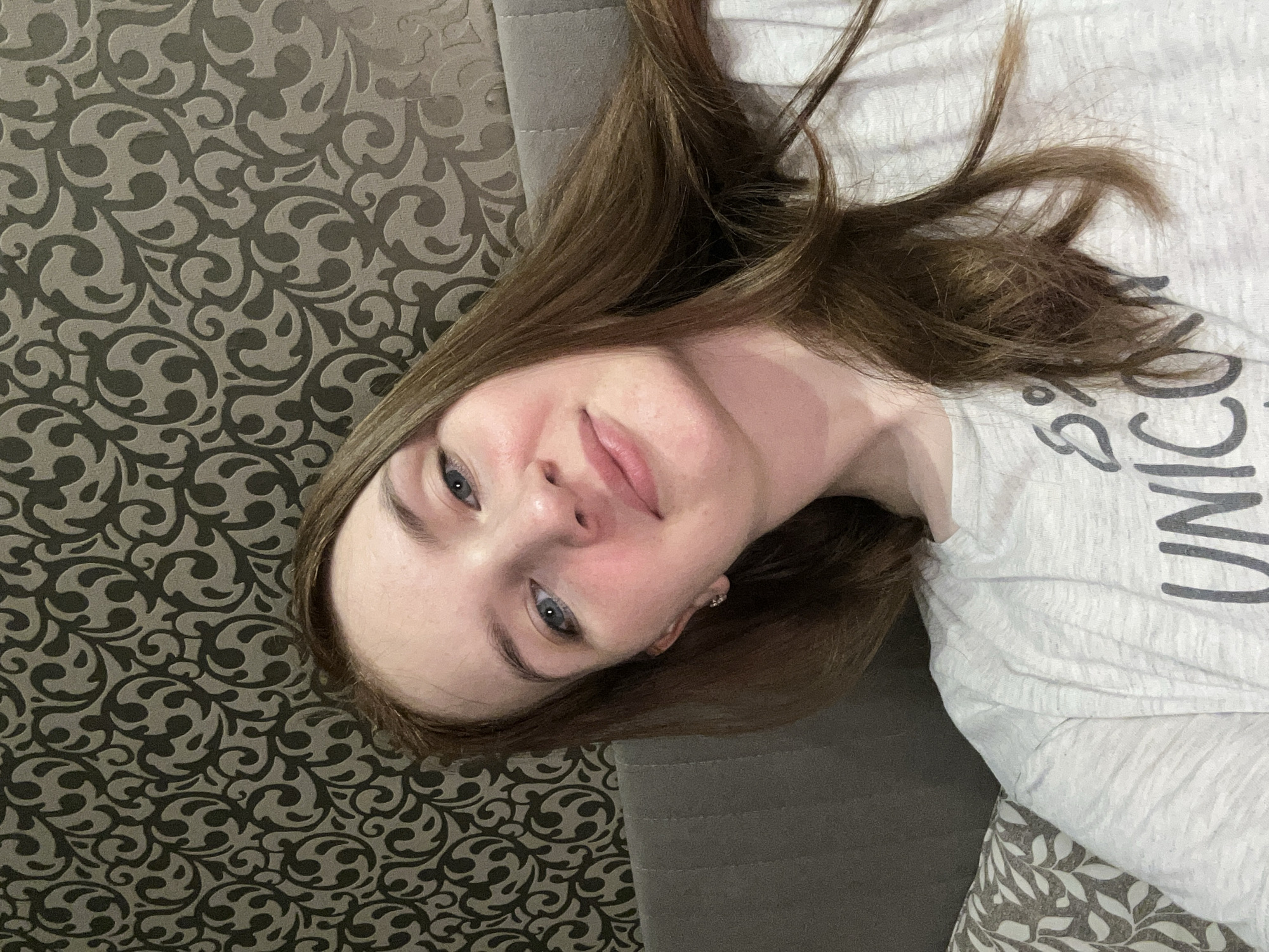 A young woman with long brown hair lies on a textured sofa in what appears to be a domestic setting. Her head is turned slightly, and she gazes at the camera with a gentle expression. She is wearing a light-colored t-shirt with what appears to be the word "UNICORN" printed on it. The sofa has a patterned dark brown and grey design. The environment consists of a patterned cushion and the couch. There is no information to indicate the location being Chaikovsky, Russia.