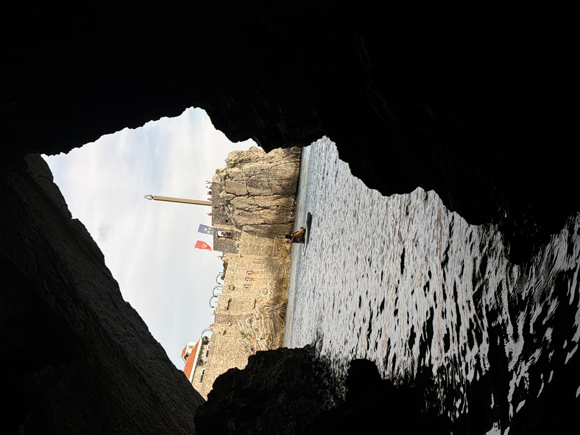 The image depicts a view from within a dark cave, looking out towards a sunlit scene of a stone fortress and the sea. The foreground is dominated by the rough, dark textures of the cave walls, which frame the opening.
Beyond the cave, a weathered stone fortress with crenellated walls and arched windows stands against a bright, overcast sky. Two flags, one red and one blue with stars, are visible flying from the fortress. A tall wooden pole extends from the top of the fortress, with what appears to be a decorative element at its apex.
In the water, a person on a paddleboard is visible near the fortress walls. The sea is relatively calm, with sunlight reflecting off its surface, creating a shimmering pattern. The water appears to be a deep blue. The overall scene suggests a historic coastal location, possibly a fortified city. The lighting indicates it is daytime, with the sun obscured by clouds. The presence of the fortress and the sea suggest a location with historical significance and natural beauty. No visible text is present in the image.