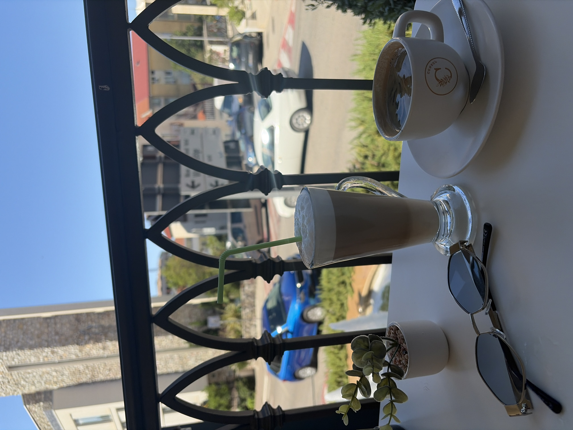 A white table is set with two drinks, sunglasses, and a small plant, on a balcony overlooking a street in Budva, Montenegro. The drinks are a tall glass of layered coffee with a green straw and a white mug of black coffee with a spoon on its saucer. The sunglasses are silver with dark lenses. The plant is in a white pot with small pebbles. In the background, cars drive on a street in front of buildings. The sky is blue.