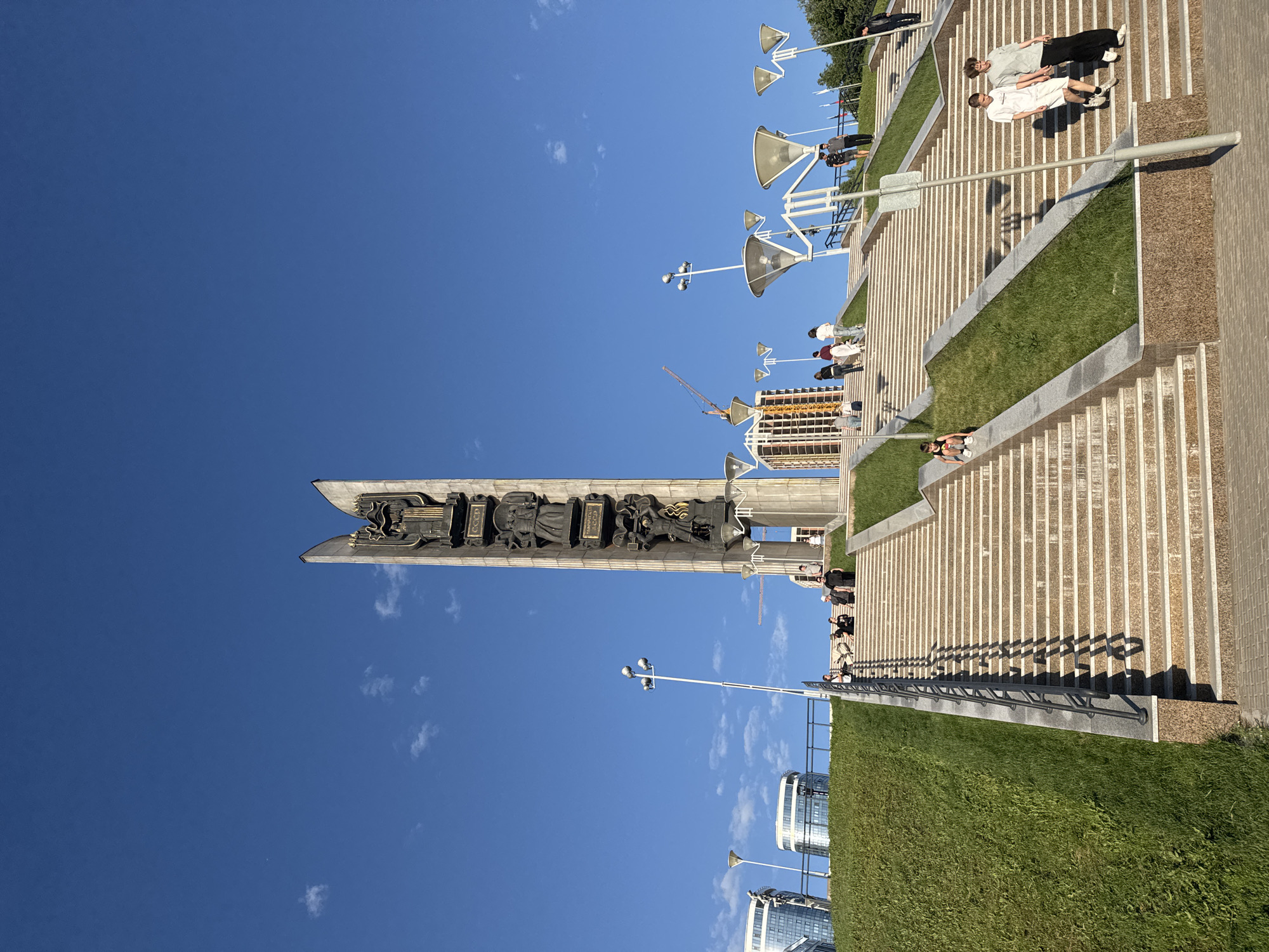 This image captures the Monument of Friendship of Peoples, also known as the Friendship Stele, in Izhevsk, Russia, under a clear blue sky with scattered white clouds. The monument, a tall, vertical stele of light-colored stone, dominates the upper center of the frame. Its front face is adorned with dark bronze relief sculptures depicting various figures, including a woman holding a torch at the apex, as well as industrial and agricultural workers, representing Soviet-era themes. The Cyrillic inscription "СССР" (USSR) is visible multiple times on the monument's façade.
In the foreground and midground, a wide, multi-tiered stairway system ascends towards the monument. The stairs are constructed from light-colored stone with darker granite accents on the risers and landings. They are divided into multiple flights by sections of neatly manicured green grass on steep embankments. Dark metal handrails with a simple design run alongside some sections of the stairs, casting long, distinct shadows on the steps due to the bright sunlight.
Various individuals are present on the stairs. On the lower right, two young men in casual attire are descending. On the lower left, a young person in a dark top and white shorts is sitting on the steps. Further up, multiple groups of people are scattered across different stair flights, some ascending, some descending, and others gathered near the monument's base.
Modern lampposts with distinctive, large, flared or bowl-shaped light fixtures are positioned along the stairs and on the upper platform. In the background to the left, several contemporary buildings with glass facades and curved architectural elements are visible atop the hill. To the right, behind the monument, a tall yellow construction crane and a building under construction can be seen. The overall scene suggests a public space or park on a sunny day.