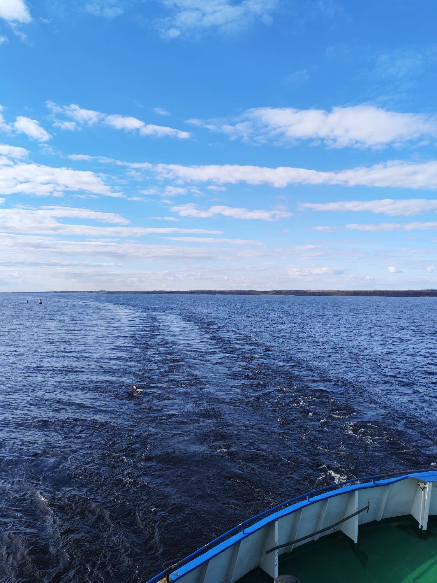 The image presents a wide, serene view from the stern of a boat moving across a vast body of dark blue water, likely a large river or lake, under a bright, partly cloudy sky. The boat's visible stern features a white hull with a prominent blue railing, and a section of green deck is visible in the bottom right corner. Faint, indistinct numbering can be seen on the white hull panel, possibly "258" or "728".
A clear, foamy wake stretches directly behind the boat, receding into the distance and indicating its forward motion. The water surface is textured with ripples and small waves. In the distance, a low, dark band of land, appearing to be a forested shoreline, extends across the horizon. A red and white navigational buoy is visible on the left side of the water, situated between the boat and the distant shore.
The sky is a brilliant blue, filled with scattered white clouds, primarily cumulus and stratocumulus types, some forming elongated bands. The weather appears fair and sunny, suggesting a pleasant day, likely morning or afternoon, given the clear visibility and bright light. No people are visible in the scene. A small, indistinct white object floats in the water near the boat's wake on the right. Based on the provided context, the location is near Shchetinskoe, Russia.