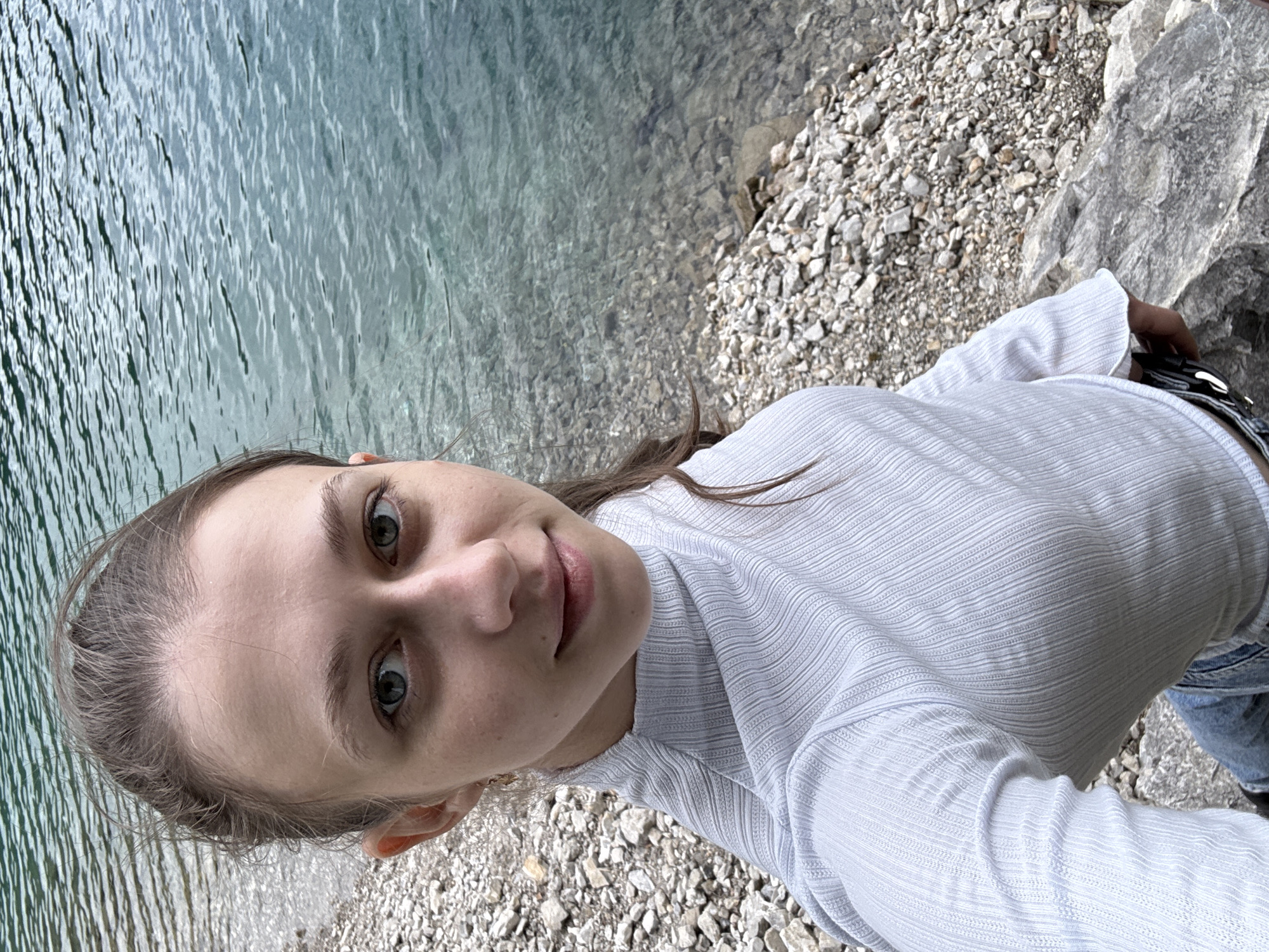 A young woman with light brown hair pulled back into a ponytail takes a selfie outdoors. She is wearing a light blue ribbed turtleneck shirt and blue jeans. She is standing on a rocky shore next to a body of clear, blue water. The water appears to be a lake or a calm river. The background shows the water and the rocky shoreline. The location is identified as Grainau, Germany.