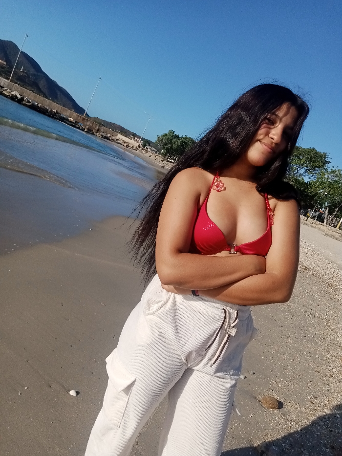 A young woman stands on a sandy beach with arms crossed. She wears a red bikini top and white cargo pants. The beach stretches to the left, meeting the ocean and a distant breakwater. A backdrop of mountains contrasts with the clear blue sky. Trees and poles are visible further down the beach to the right. Sunlight illuminates the scene, casting light and shadows on the sand.
