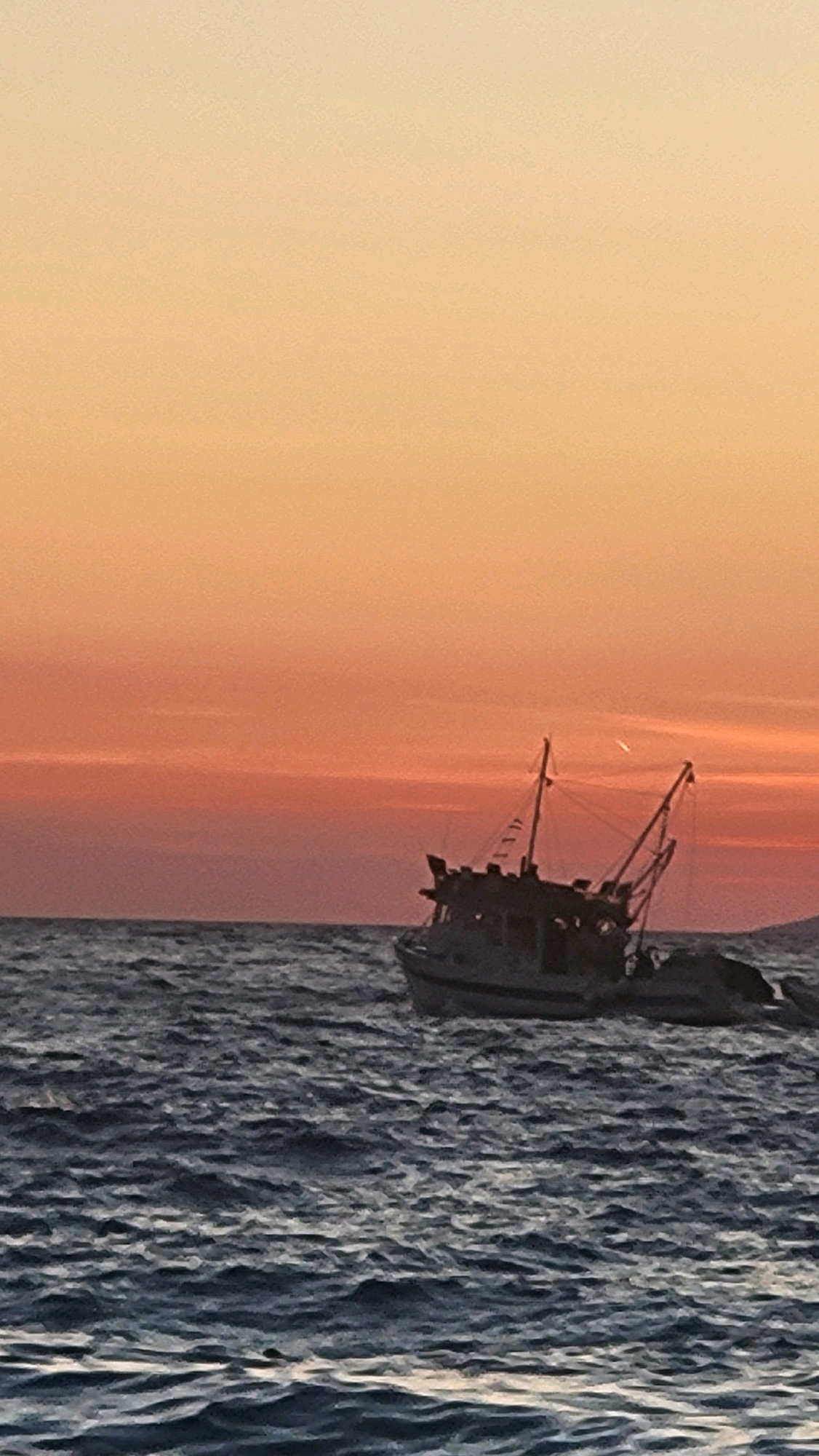 A fishing boat is navigating the choppy waters of the Adriatic Sea off the coast of Općina Tučepi, Croatia, at sunset. The sky is a gradient of soft orange and pink hues, with a faint contrail visible high above. The boat, a medium-sized vessel with a dark hull and a visible superstructure, is silhouetted against the vibrant sky. The sea surface is textured with numerous waves and whitecaps, reflecting the dimming light. The scene conveys a sense of quiet activity and the end of a day at sea.
