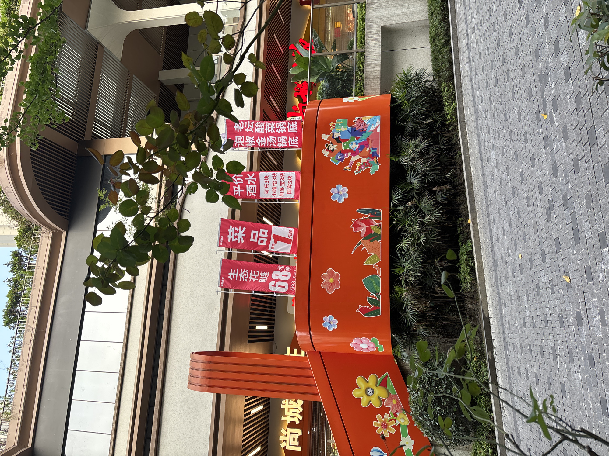 The image captures a vibrant street scene in Nan'an District, China. A large, orange decorative structure stands prominently, adorned with cartoonish figures and floral designs. Banners displaying Chinese characters and promotional offers are visible. Buildings with architectural details are in the background, with foliage partially obscuring the view. A paved walkway extends along the side, bordered by greenery.