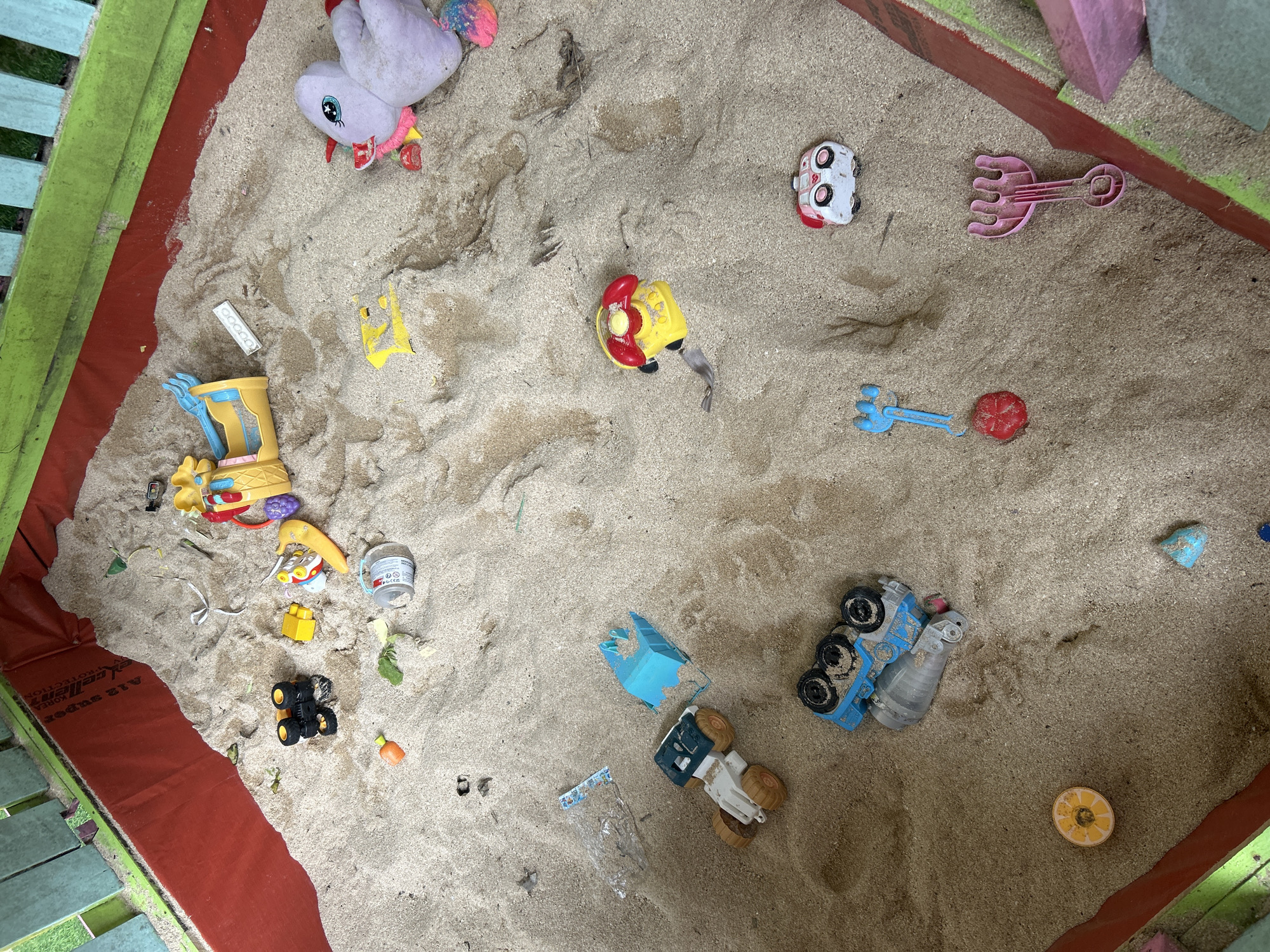 The image shows a sandbox filled with various toys, set within a fenced outdoor area. The sandbox has a red liner and is surrounded by green and light blue painted wooden fencing. Among the toys scattered in the sand are a purple stuffed animal with a rainbow-colored tail, a yellow toy telephone, a toy truck with a blue and gray body, a blue toy shovel, and several smaller toys including a red circular object, a yellow toy car, and a small white toy vehicle with red markings. There are also some green leaves and other debris mixed in with the sand. The scene appears to be a child's play area. The location is identified as Kuta, Indonesia.