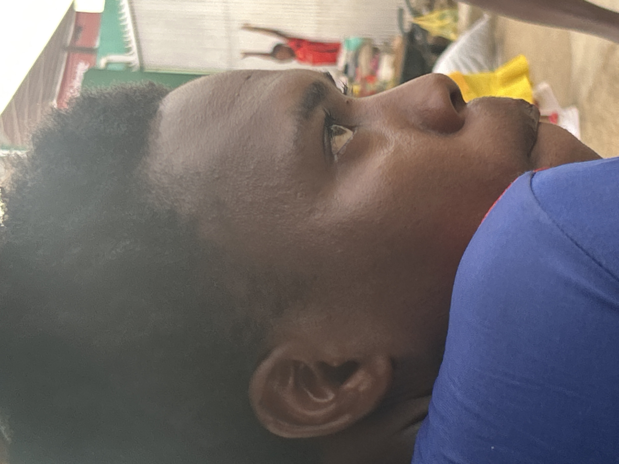 An eye-level close-up shot shows the side profile of a dark-skinned person wearing a blue shirt. The person has short black hair and a visible ear. The background is blurry, but appears to be an outdoor scene, including another person that looks like a poster behind the person in the blue shirt wearing a red shirt, with a yellow object visible as well. While the image offers no concrete evidence of its location, there is no verifiable connection of this image to Jos, Nigeria.