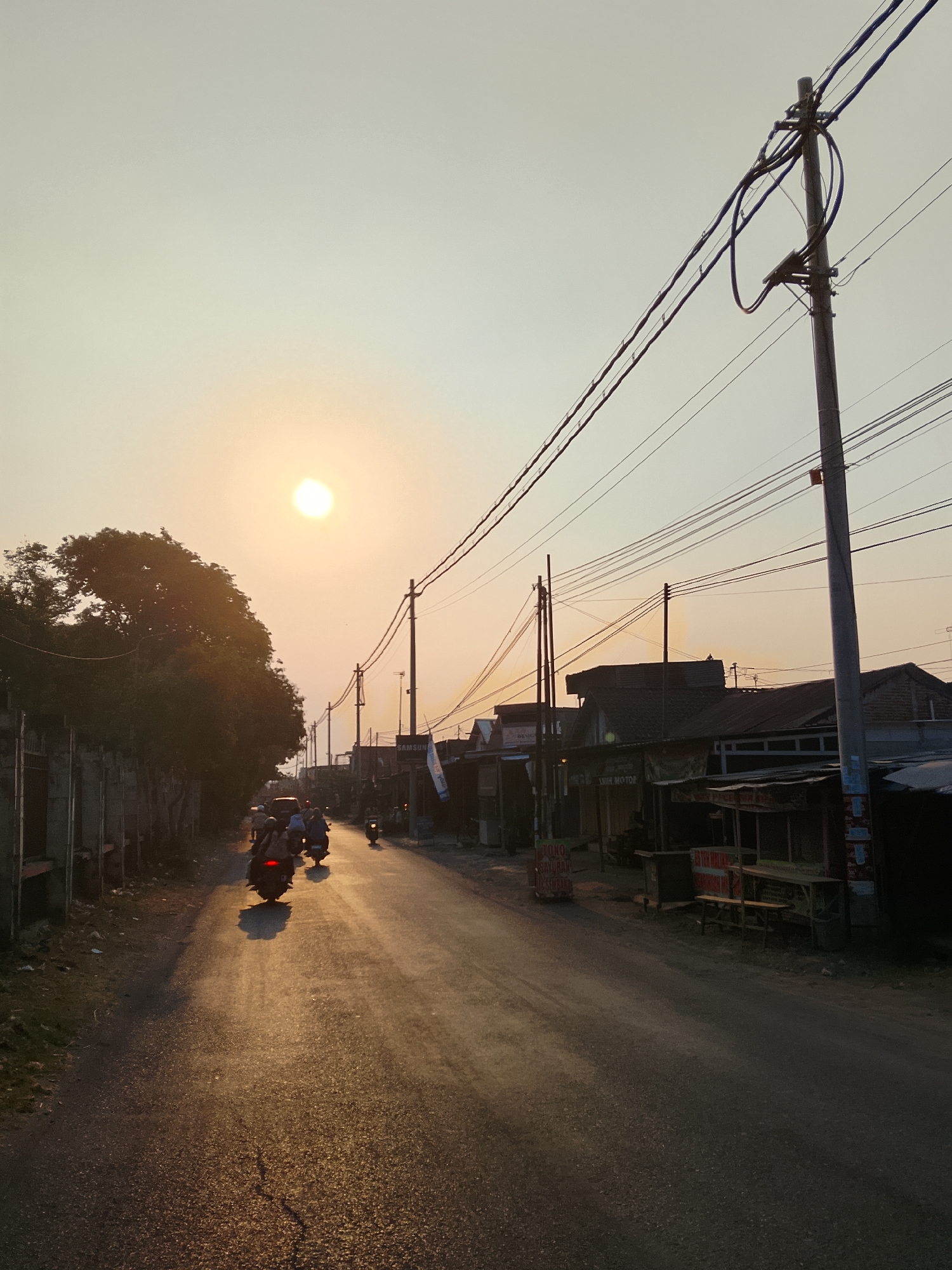 This is a street-level view of Ngelo, Indonesia, captured during the golden hour of sunset. The scene depicts a paved road lined with modest buildings and overhead electrical wires supported by wooden poles. Several individuals are on motorcycles, traveling along the road, casting long shadows. The sun, a bright orb, is low in the sky, casting a warm, orange glow over the landscape. On the right side of the road, there are small storefronts or market stalls, some appearing to be selling goods like vegetables. The overall atmosphere is one of a bustling, everyday scene in a residential or commercial area of Indonesia during dusk.