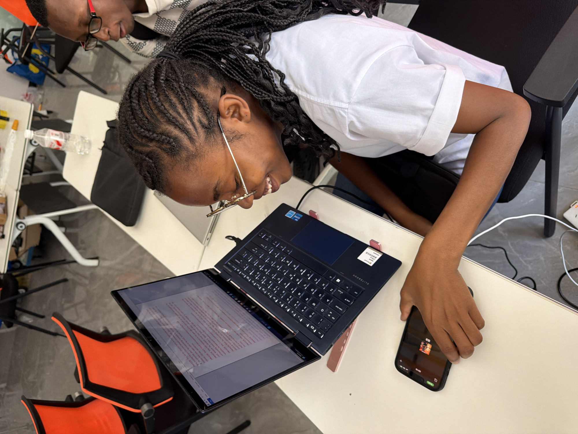 In a room resembling an office or classroom in Jiangjin, China, a young Black female student is seated at a white table, engrossed in her laptop. Her braided hair is pulled back in a ponytail, and she wears glasses and a white t-shirt. She is smiling and looking at her laptop, which is open and displaying text. Her right hand rests on the table, near a smartphone. To the right of the student, another Black individual, wearing glasses and a patterned shirt, is partially visible, also looking towards the laptop. In the background, orange chairs are arranged around other tables. The environment appears to be an indoor workspace with various office supplies and equipment visible.