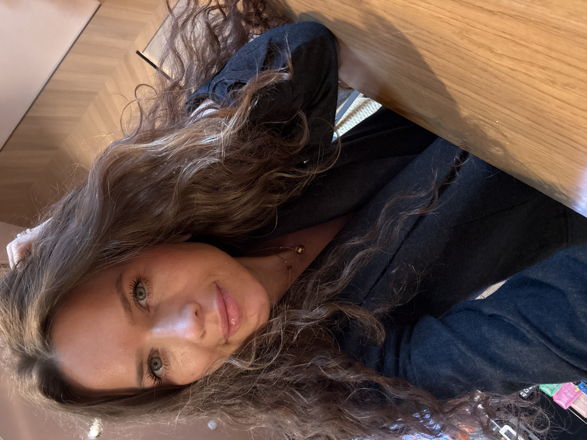 A woman with long, wavy brown hair and blue eyes is shown in a selfie. She is wearing a black top and a necklace with a gold pendant. The background appears to be an indoor setting with a wooden table and a parquet floor. There is no visible indication of this location being in Moscow, Russia.
