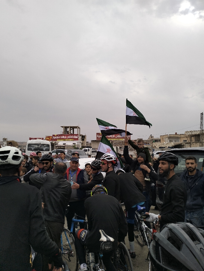 A group of cyclists and onlookers gather in a street, some holding Syrian flags. The cyclists are wearing helmets and cycling gear, while the other people are dressed in casual clothing. A man holds a child on his shoulders. The scene appears to be a celebration or rally, with people interacting and possibly shaking hands. Vehicles are parked nearby, and buildings are visible in the background under an overcast sky.