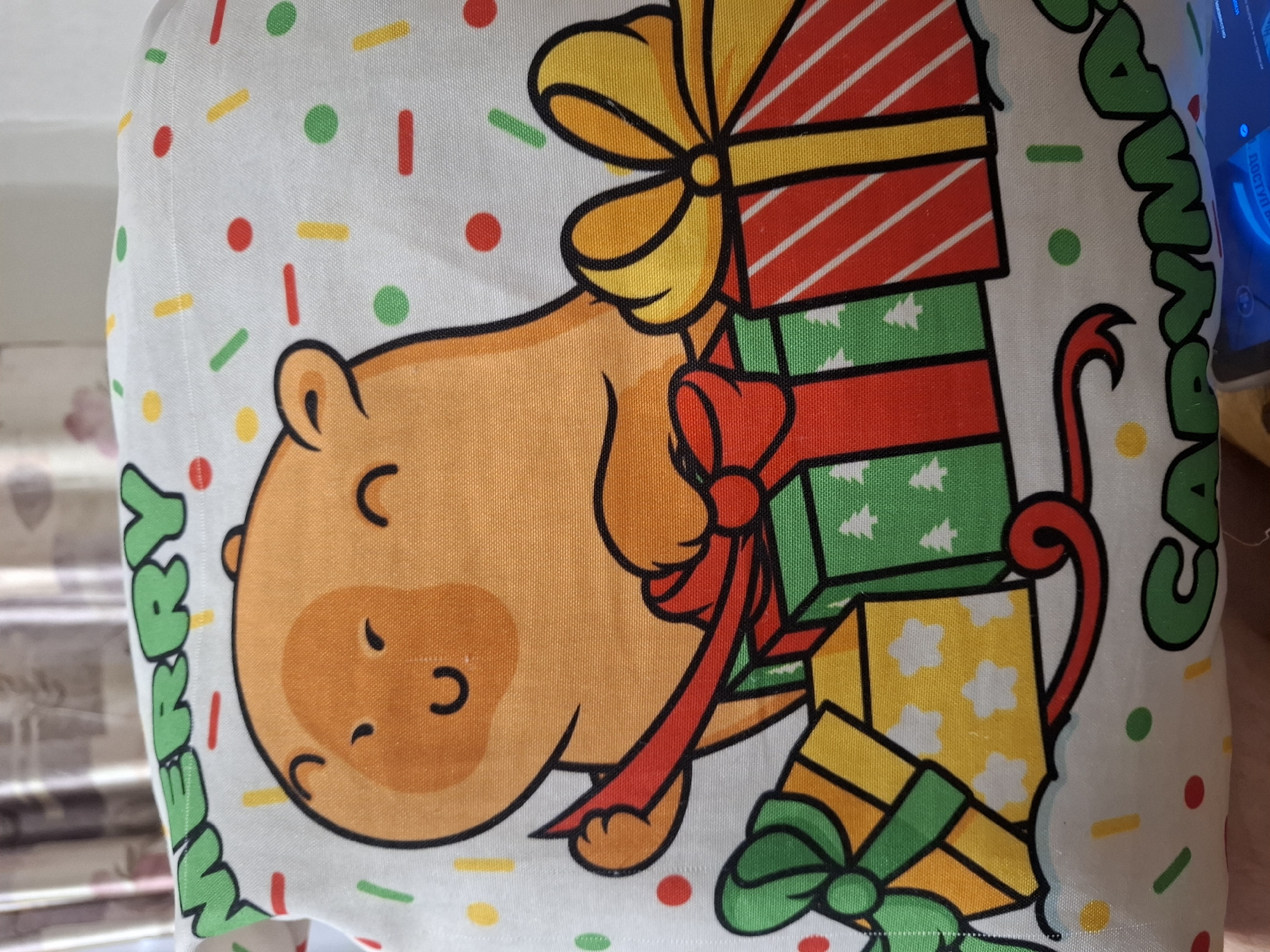 The image shows a close-up of a white fabric with a Christmas-themed cartoon print. The print features a happy-looking orange capybara character surrounded by gift boxes. The capybara is giving a thumbs-up gesture. The fabric is decorated with colorful confetti dots and lines in red, green, yellow, and white. Text reading "MERRY" in green letters is visible at the top, and partially visible text at the bottom appears to say "CAPYMAS". The fabric appears to be part of a pillow or a similar decorative item. The background shows blurred curtains. There is no identifiable location in the city of Novlyanka, Russia, visible in this image.