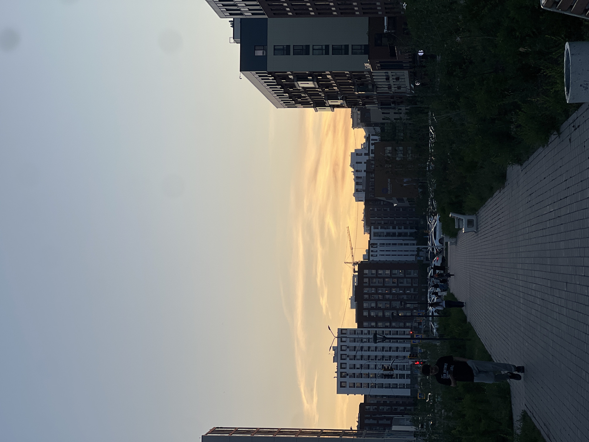 This is a photograph taken at dusk in Astana, Kazakhstan, likely in a residential complex. In the foreground, a paved walkway stretches into the distance, bordered by dense, dark green foliage on the right and a strip of grass on the left. A young man in a black t-shirt and gray pants walks away from the viewer on the walkway. Further down the path, several other people are walking, and a white bench is visible. A parking lot filled with cars is situated behind the walkway and foliage.
The background is dominated by a cluster of modern, multi-story apartment buildings. The architecture varies, with some buildings featuring light-colored facades and numerous windows, while others are darker. A construction crane is visible in the distance, suggesting ongoing development. The sky above is a soft gradient of pale blue and warm orange hues, indicating the setting sun. The overall atmosphere is calm and urban.