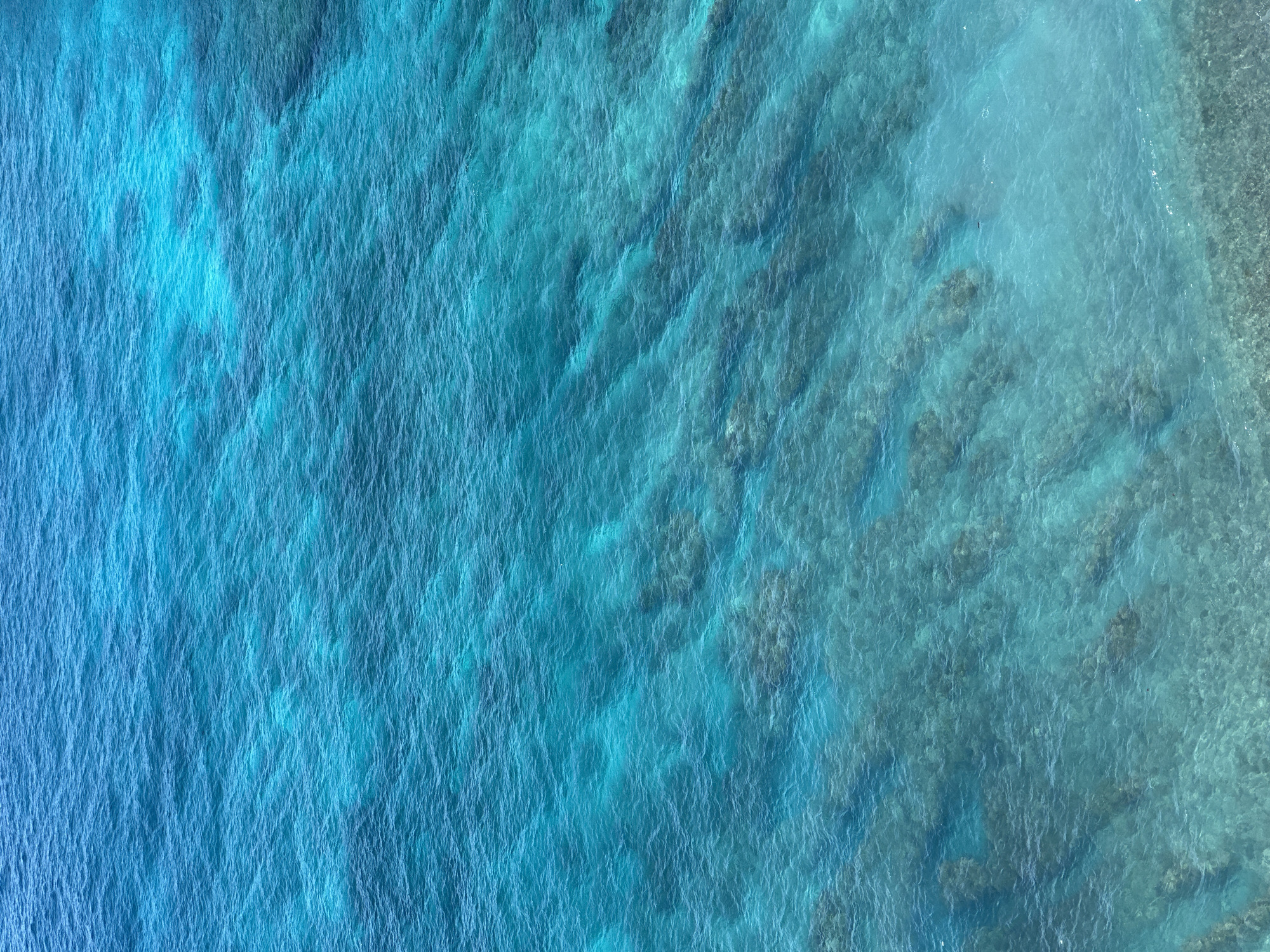 This aerial photograph depicts the ocean surface near Nusa Penida, Indonesia. The water is predominantly a gradient of blues, transitioning from a deeper indigo on the left to a lighter turquoise towards the right. Submerged coral reefs and rocks are visible through the clear water, giving the ocean floor a textured appearance. The surface of the water exhibits patterns of subtle ripples and waves, indicating movement. Along the right edge of the image, the water appears shallowest, transitioning to a lighter, almost transparent color, suggesting proximity to the shore.