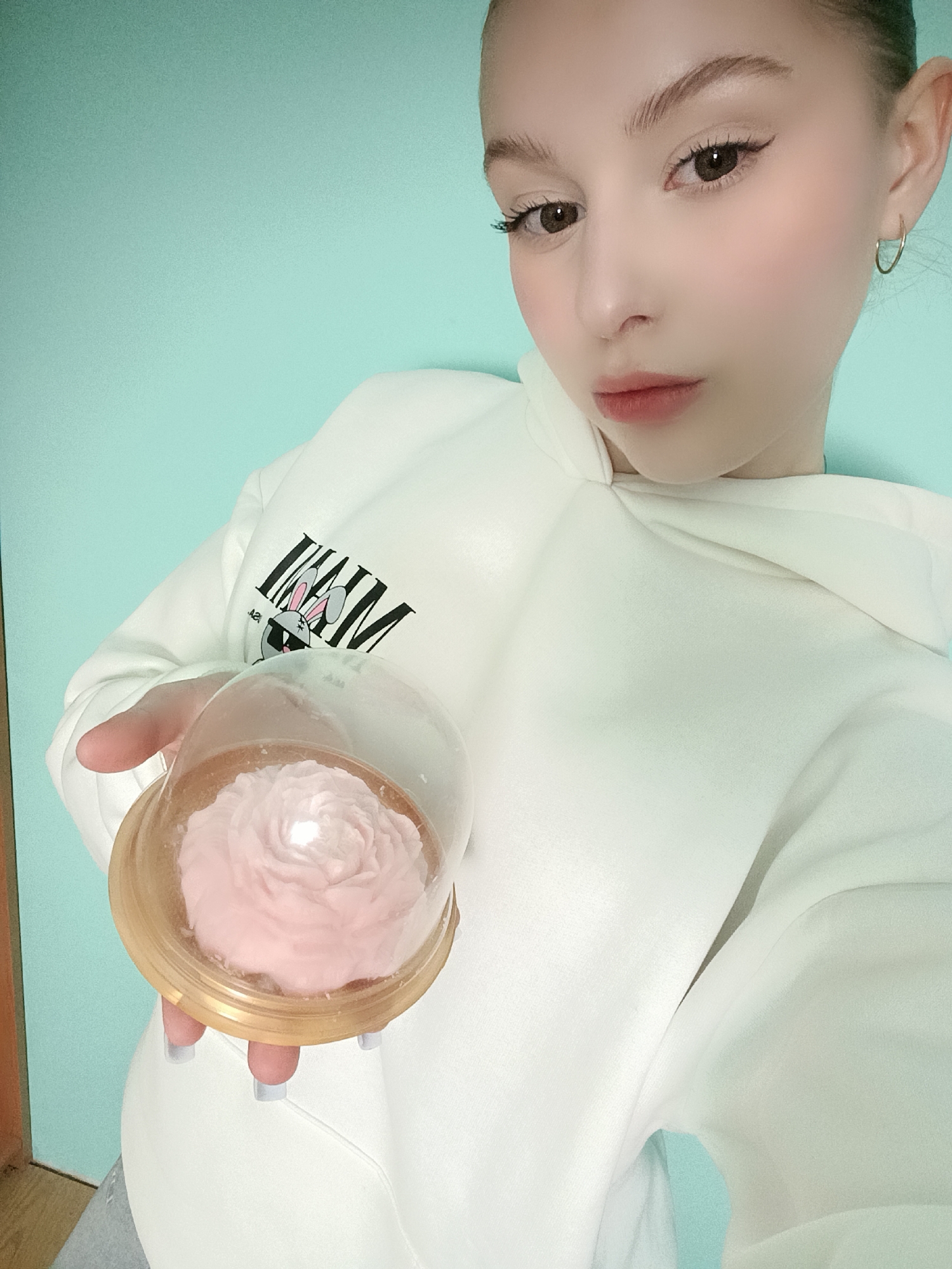 A young woman is holding a rose-shaped cake in a clear plastic container. The container has a gold-colored base. She is wearing a white hoodie with a cartoon rabbit design on the front. Her nails are painted light grey. She has light skin, brown eyes, and her hair is pulled back. She is wearing small hoop earrings. The background is a light blue wall. There is a wooden doorframe visible on the left side of the image. It's not possible to confirm the location as Chaikovsky, Russia, or any other specific location based solely on the visual information provided.